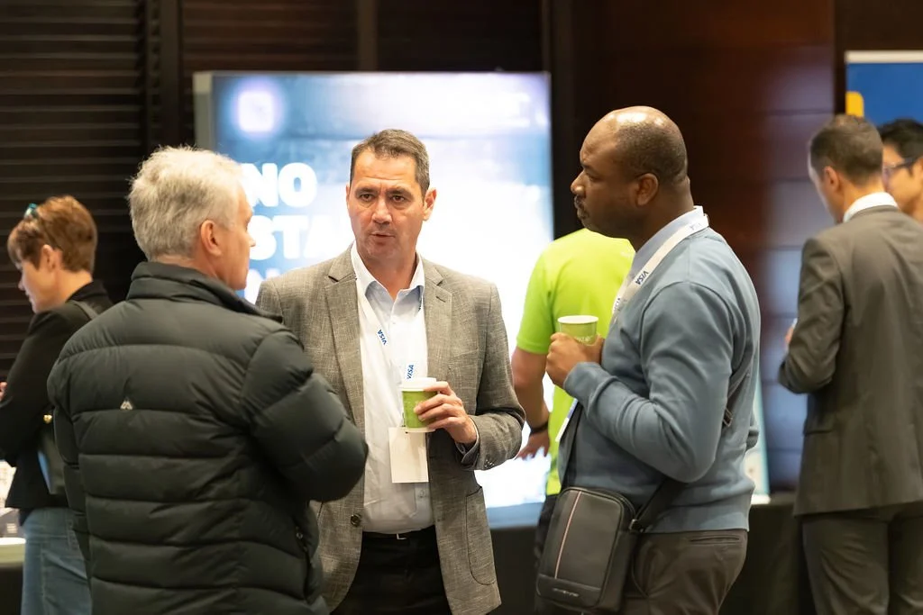 Three men engaged in conversation at a professional event, each holding a green paper cup. Additional people are visible in the background, with a screen displaying text behind them.