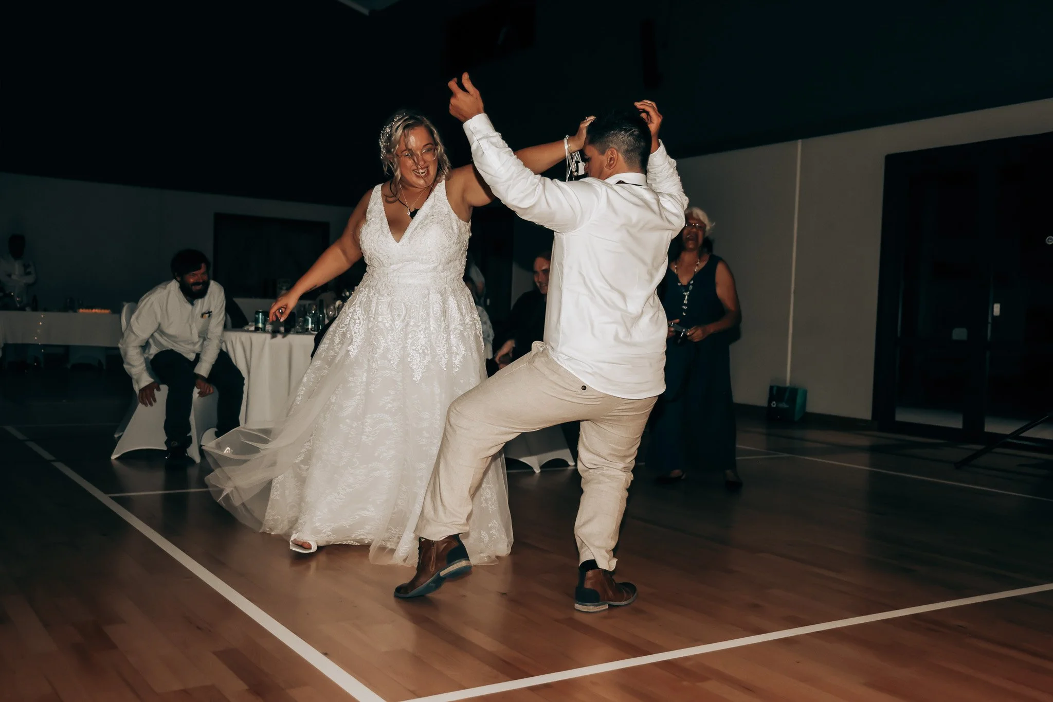 A bride and groom are dancing together at a wedding reception, smiling and enjoying the moment, with seated guests and a table in the background.