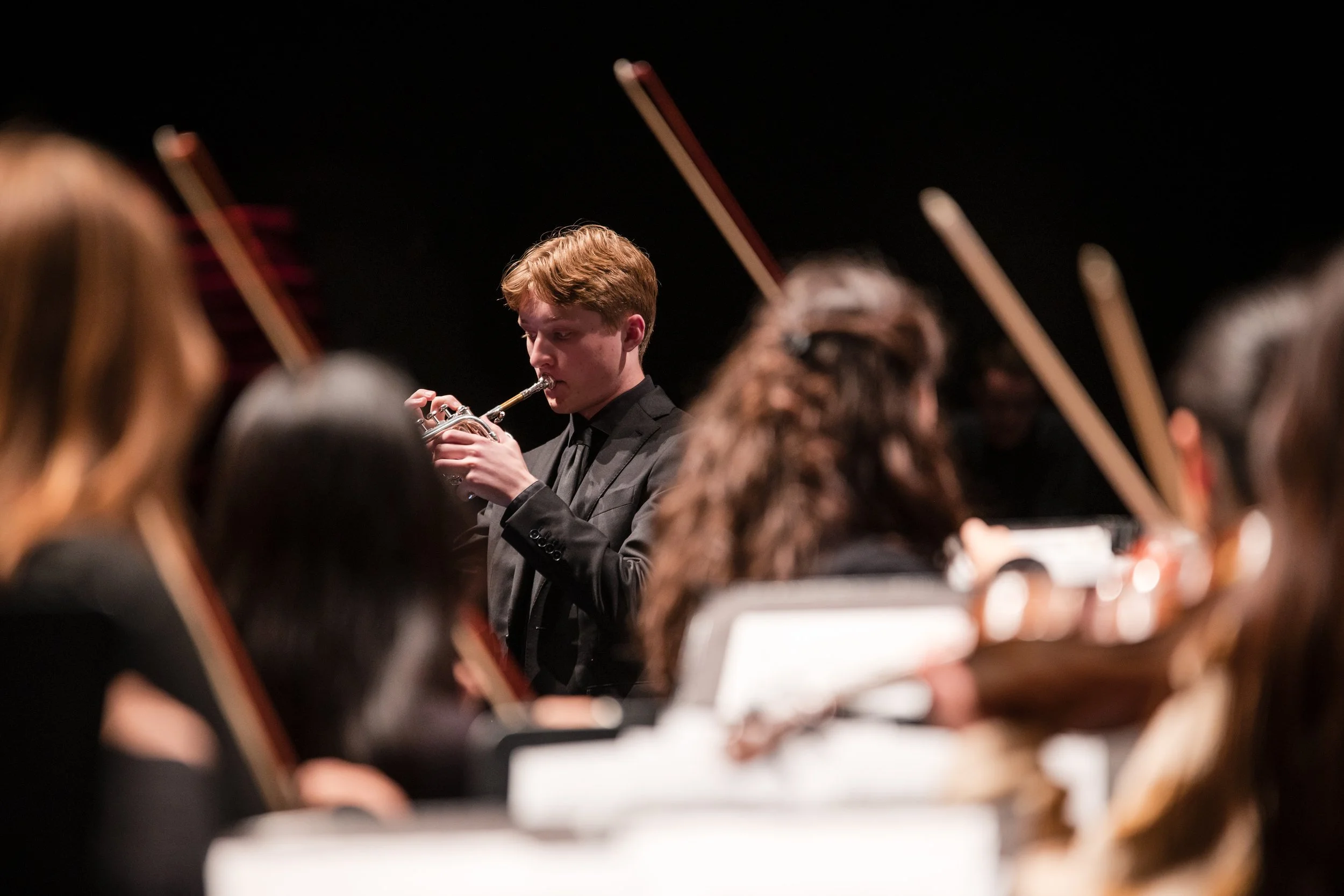 A young man in a black suit playing the trumpet in an orchestra, with other orchestra members visible and blurred in the foreground.