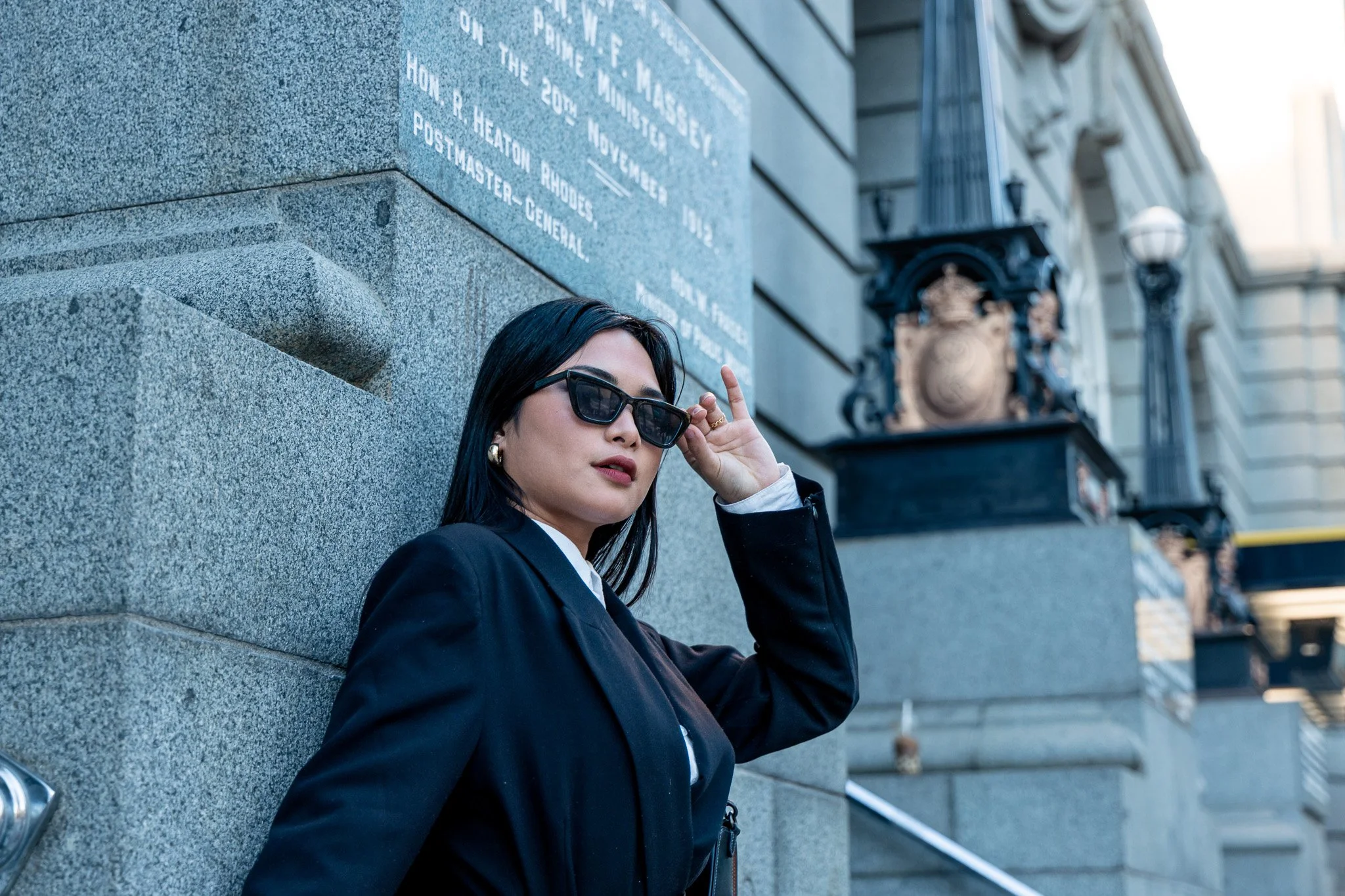 A woman with black hair, sunglasses, and earrings is leaning against a stone wall in an urban setting, adjusting her sunglasses with her right hand, with city buildings and street lamps in the background.