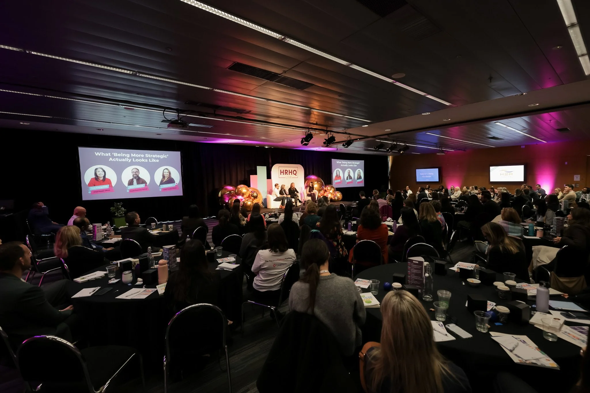 A conference room filled with people seated at round tables, listening to a panel of speakers on stage. There are large screens displaying presentation slides, and purple lighting accentuates the room's decor.