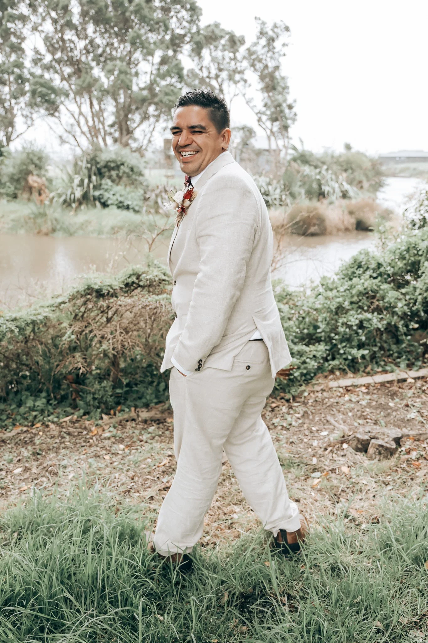 A man in a beige suit smiling and walking outdoors near a river, surrounded by greenery and trees.