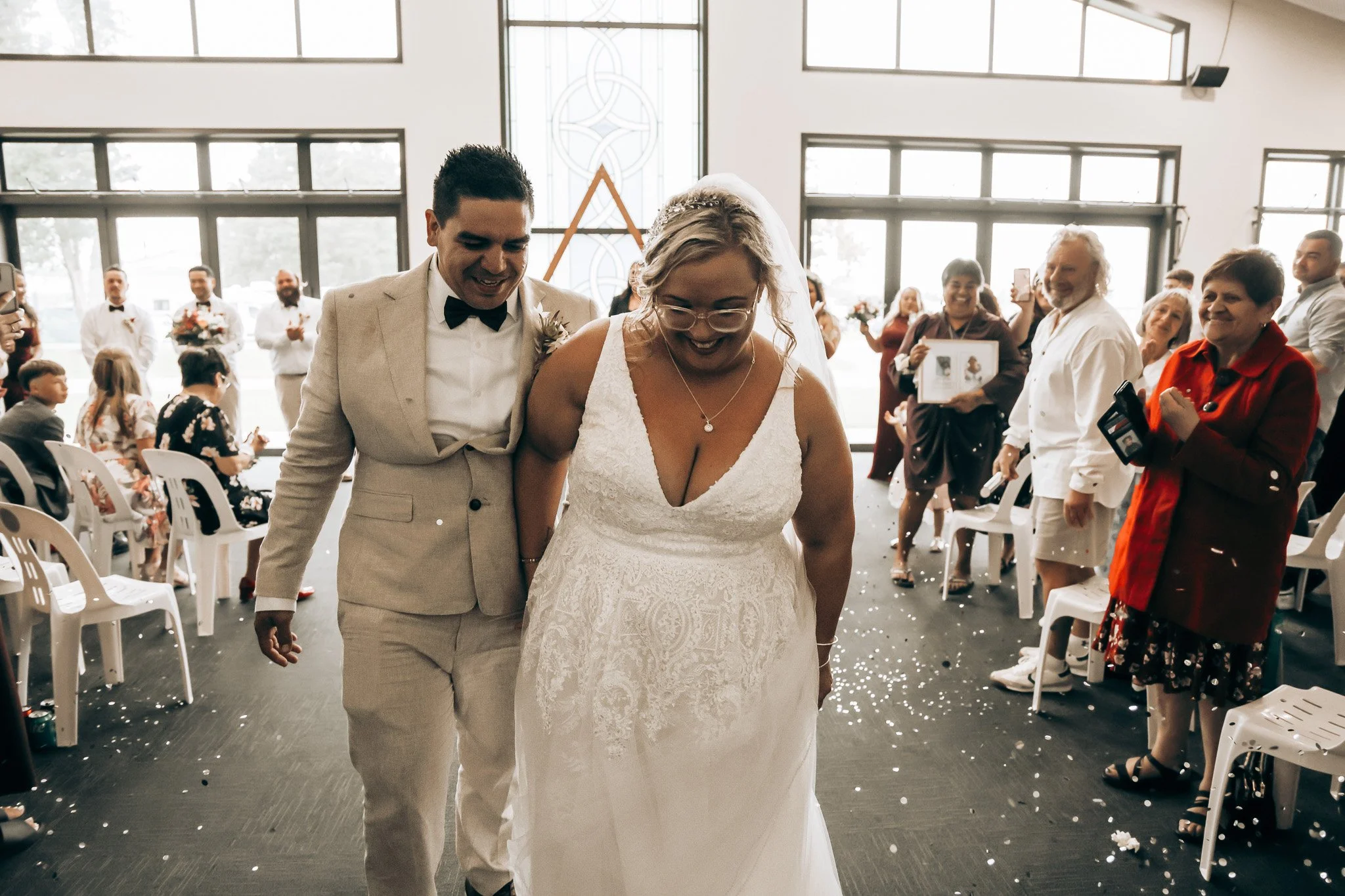A bride and groom walking down the aisle at a wedding ceremony, surrounded by smiling guests clapping and taking photos inside a bright, modern venue with large windows and seating on both sides.