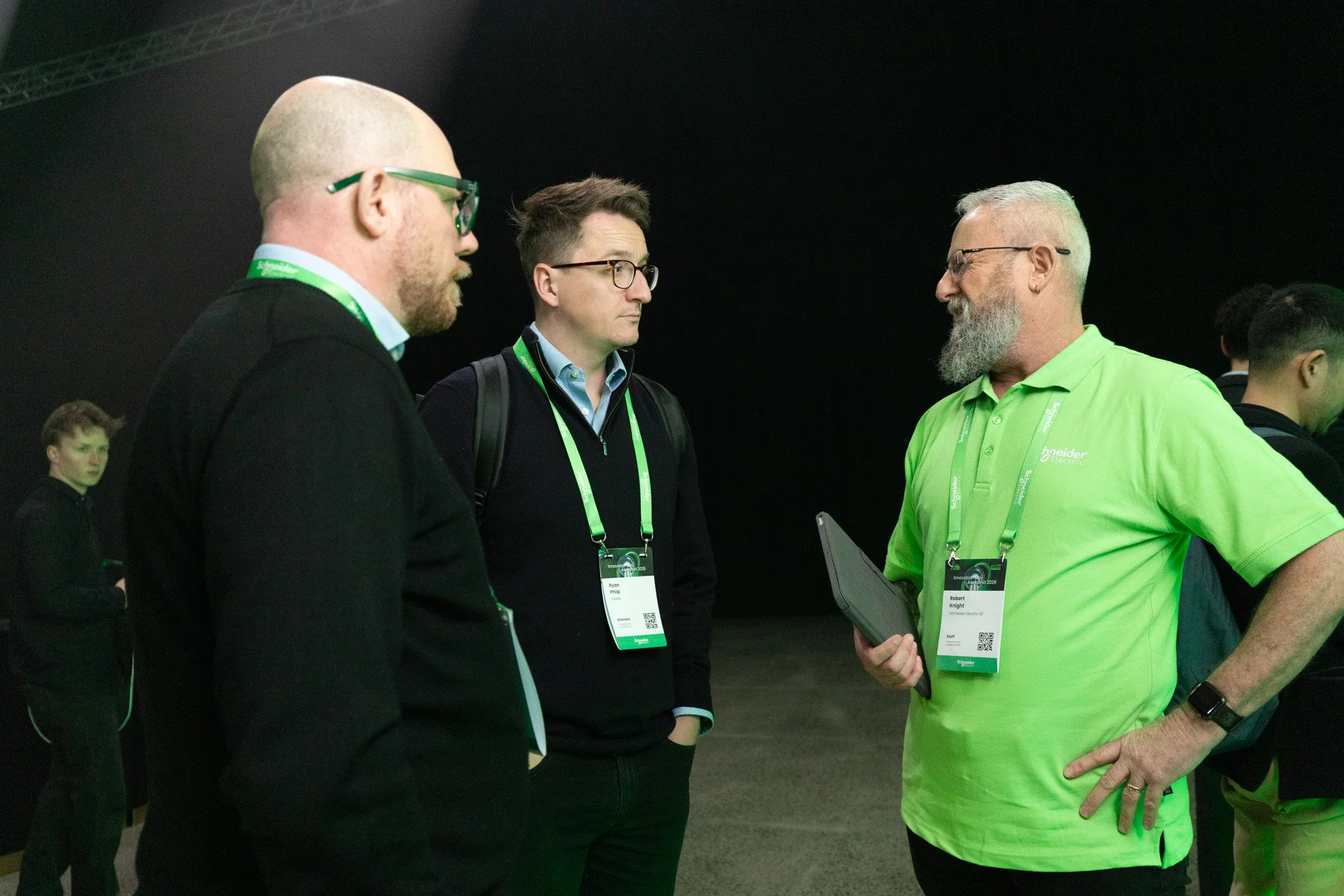 Three men engaged in conversation at a professional event, wearing conference badges with lanyards. The man on the right, dressed in a bright green shirt, appears to be showing something to the other two men, who are listening attentively.