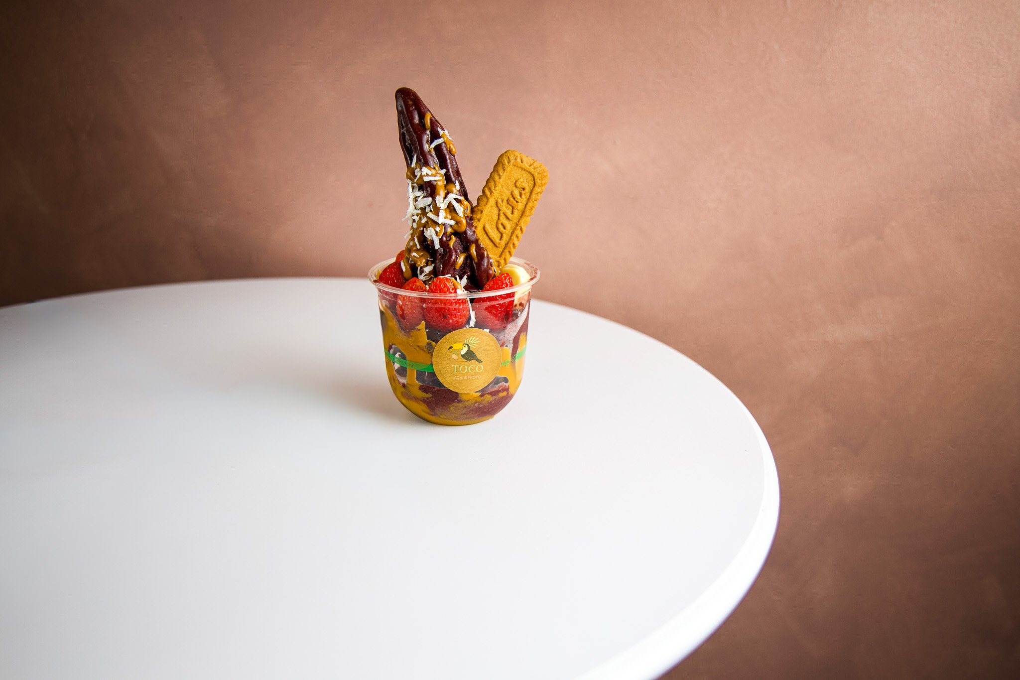 A cup with strawberries, a chocolate-coated ice cream bar, a biscuit, and coconut flakes on top, placed on a white round table against a brown background.