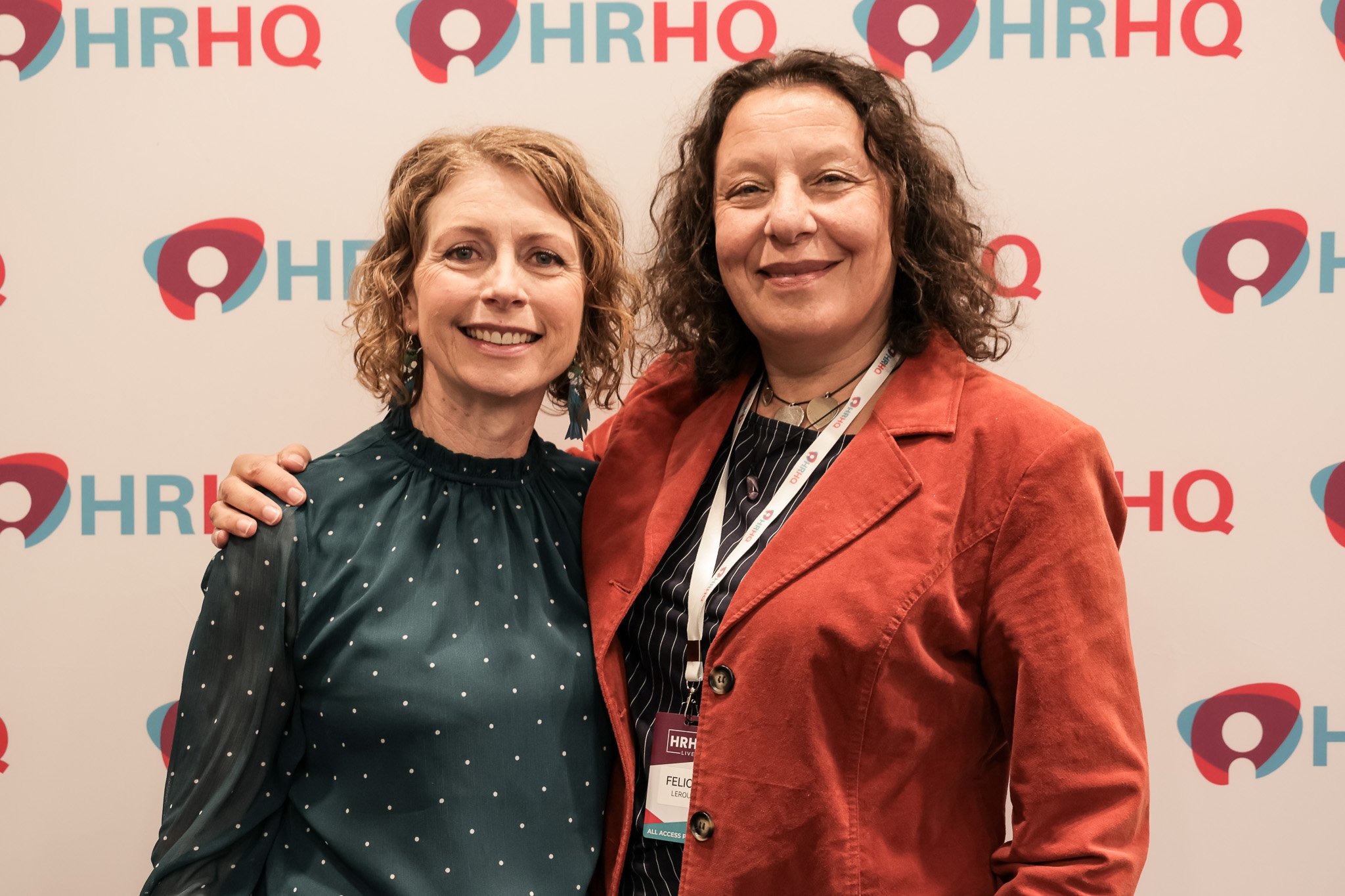 Two women standing close together in front of a white backdrop with the HRHQ logo repeatedly printed in red and teal. The woman on the left has curly blonde hair, is smiling, and is wearing a dark green dress with white polka dots. The woman on the r