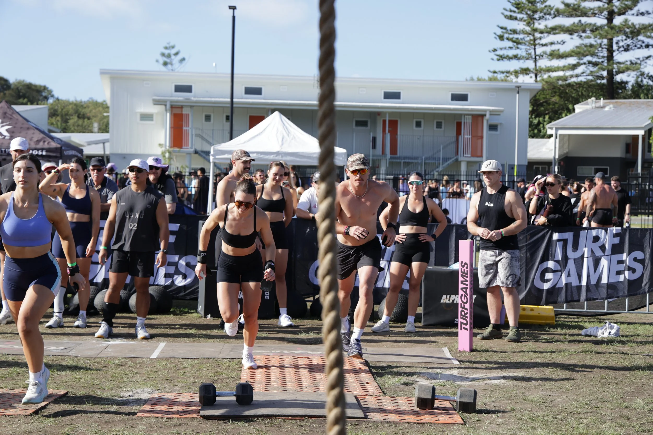 Group of athletes in athletic wear preparing for a race outdoors, with some stretching and others standing near a starting line, tents, and trees in the background.