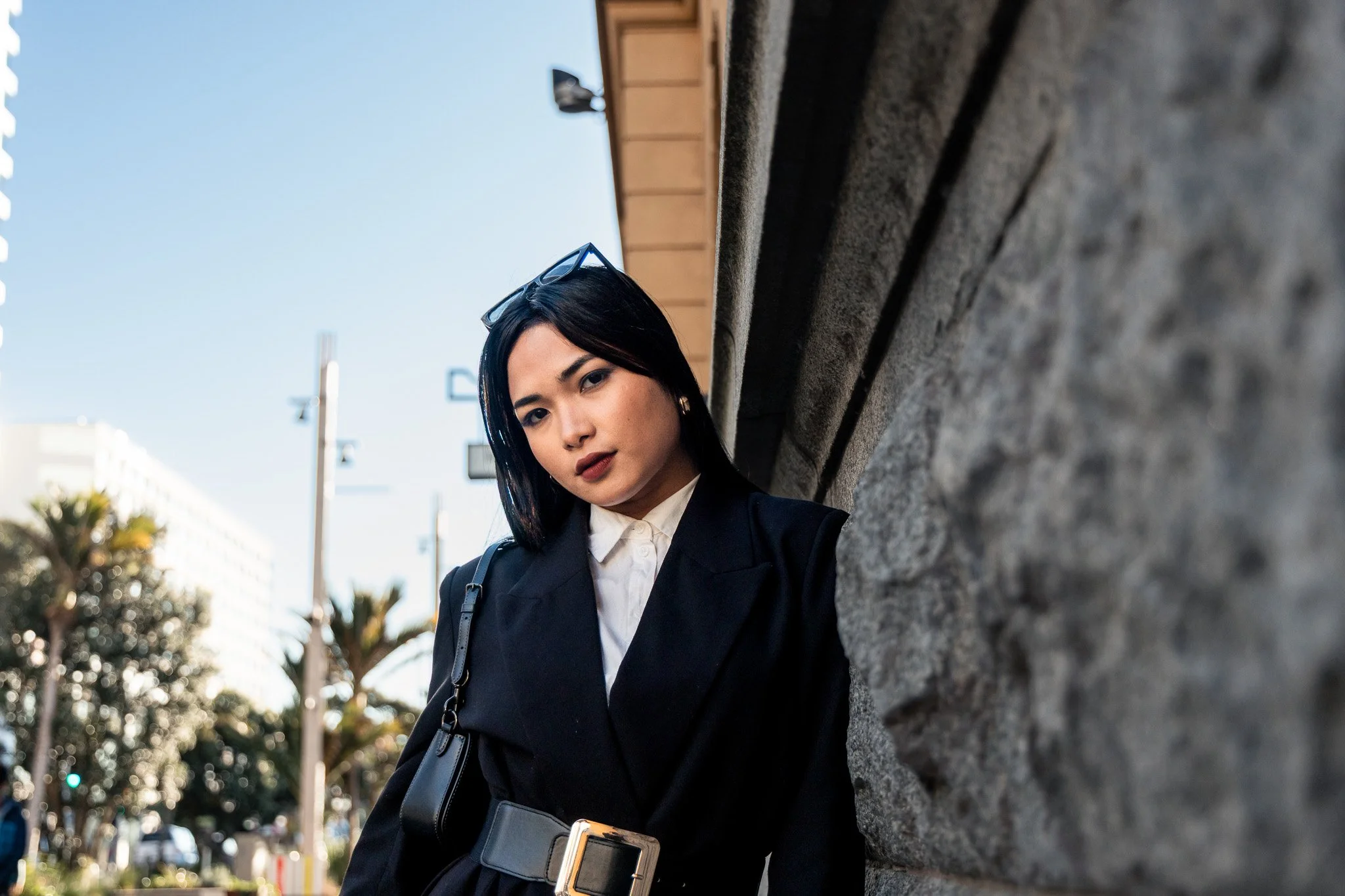 A young woman with dark hair and makeup, wearing glasses, a white collared shirt, and a black blazer, leans against a brick wall outdoors on a sunny day with a city street, trees, and buildings in the background.