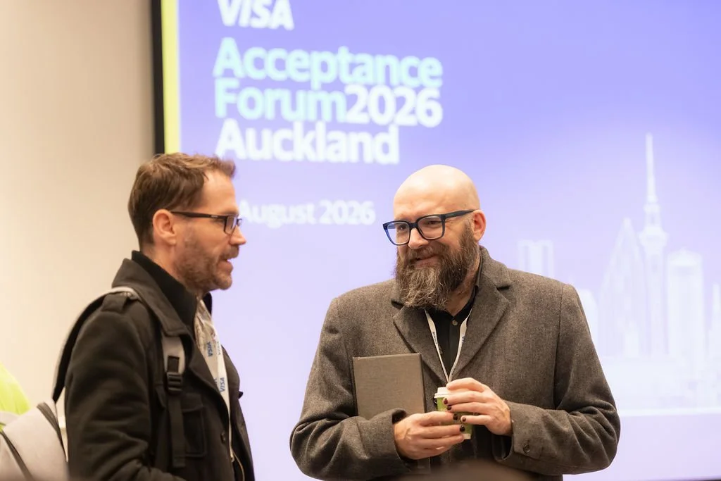 Two men talking at the Visa Acceptance Forum 2026 in Auckland, with a large screen behind them displaying event details and city skyline.