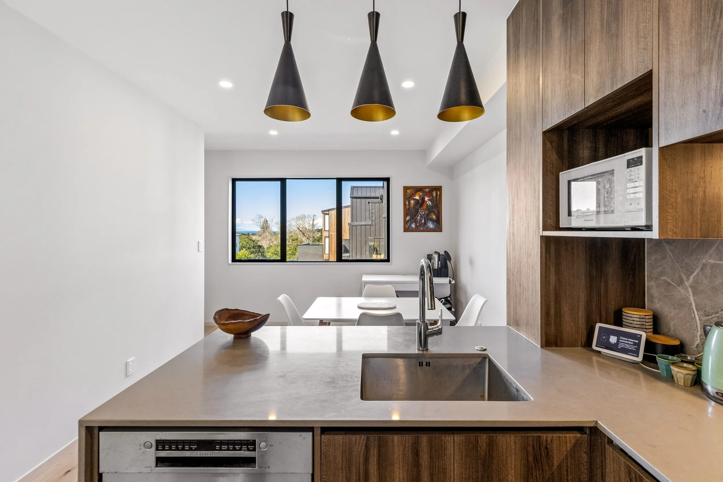 A modern kitchen with a white wall, pendant lights, a window with a view of trees and neighboring buildings, and a microwave built into wooden cabinetry.