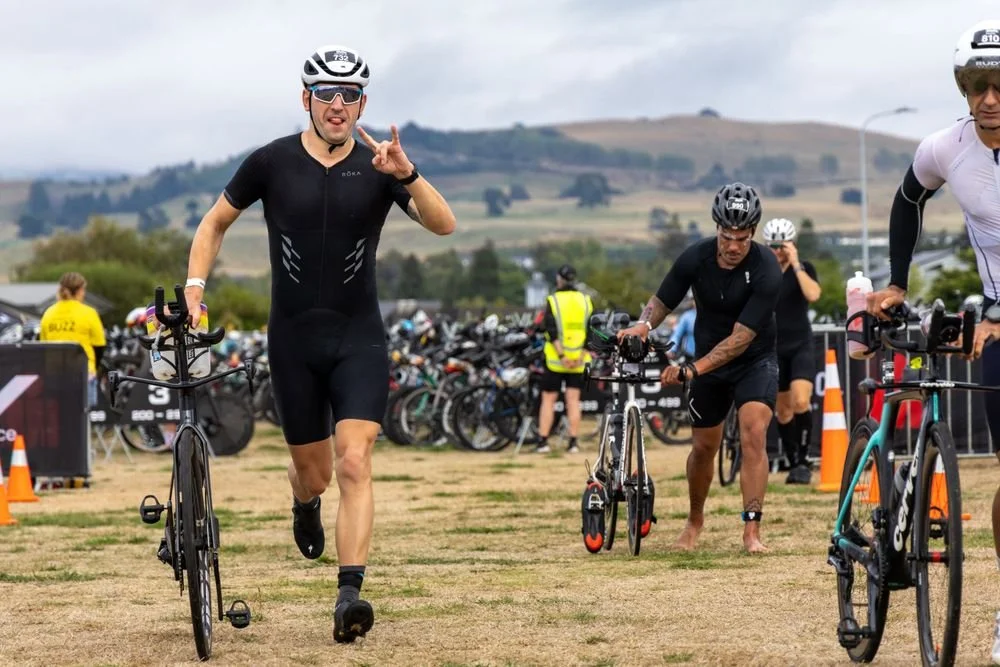 Triathlon athletes with bicycles at the transition area, some running, others adjusting their bikes, with a backdrop of hills and overcast sky.