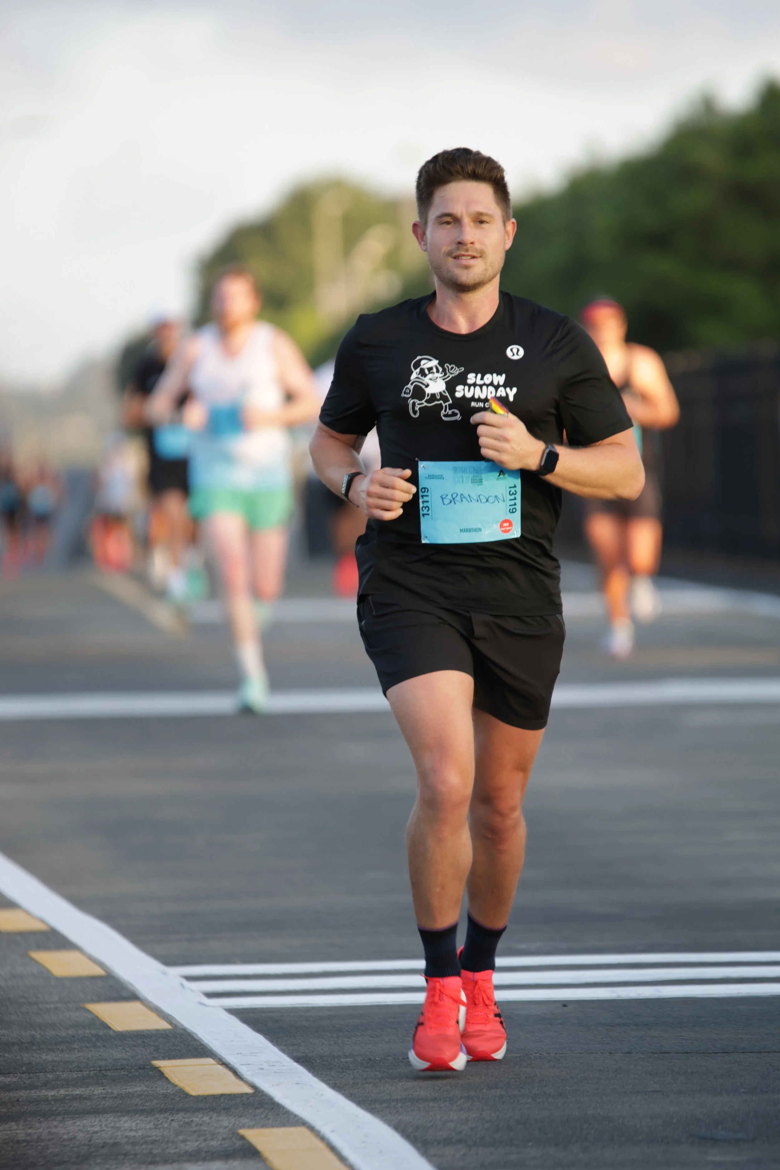 A man running in a marathon, wearing a black shirt with a cartoon character and the phrase 'Slow Sunday' on it, black shorts, red running shoes, and a watch, with others running behind him on a paved road with white and yellow markings, trees in the 