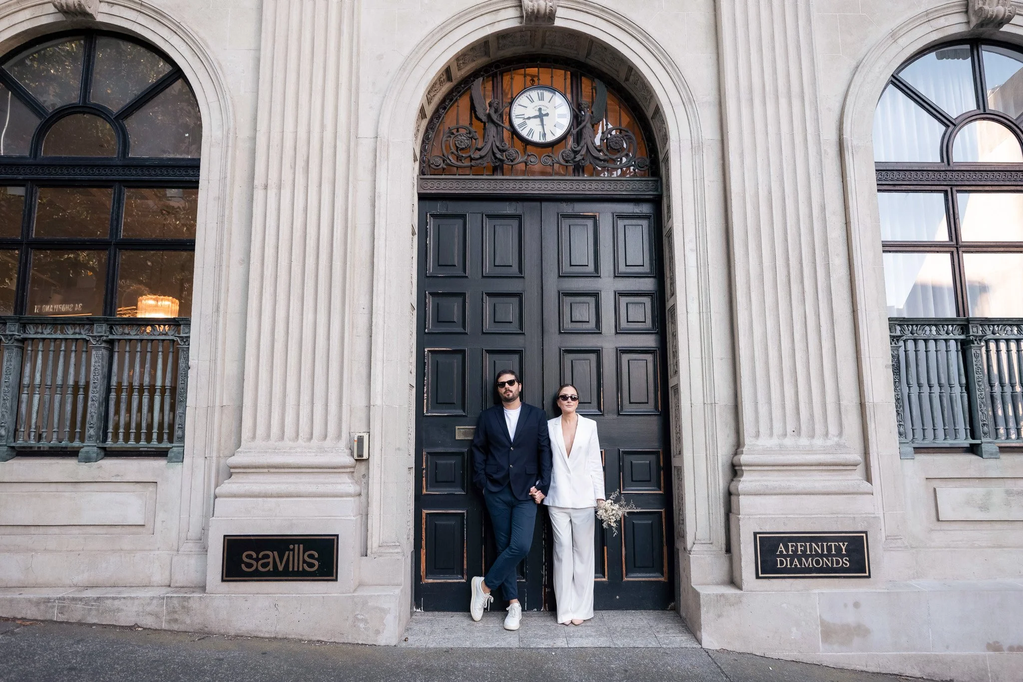A man and woman stand in front of a large black wooden door with a clock and decorative ironwork above it, set in a stone building with arched windows and columns, with two plaques reading 'savills' and 'AFFINITY DIAMONDS'. the man is wearing a dark 