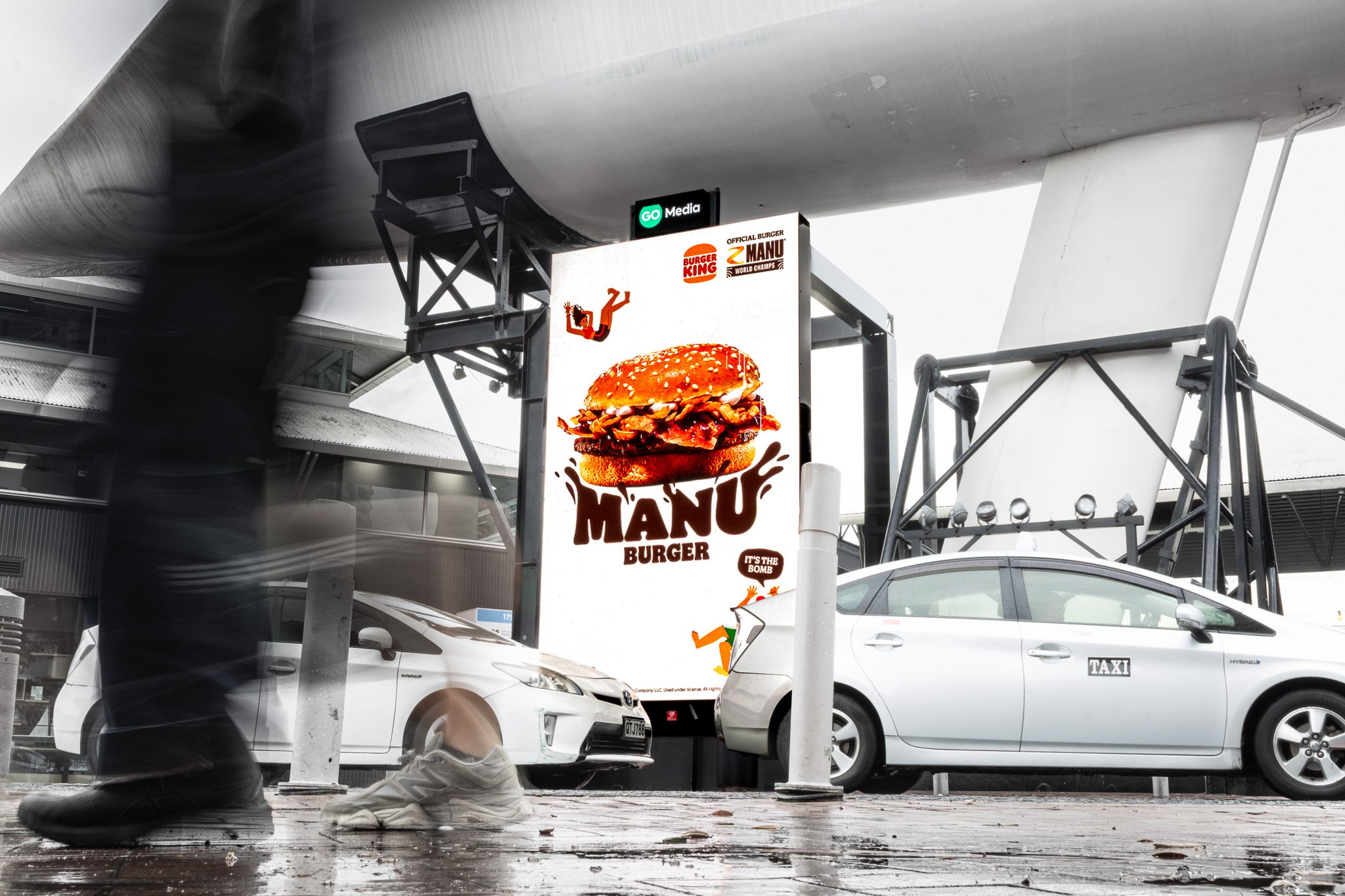 A large illuminated billboard advertising a burger called 'Manu Burger' with a picture of a burger, in a parking lot with cars and pedestrians passing by.