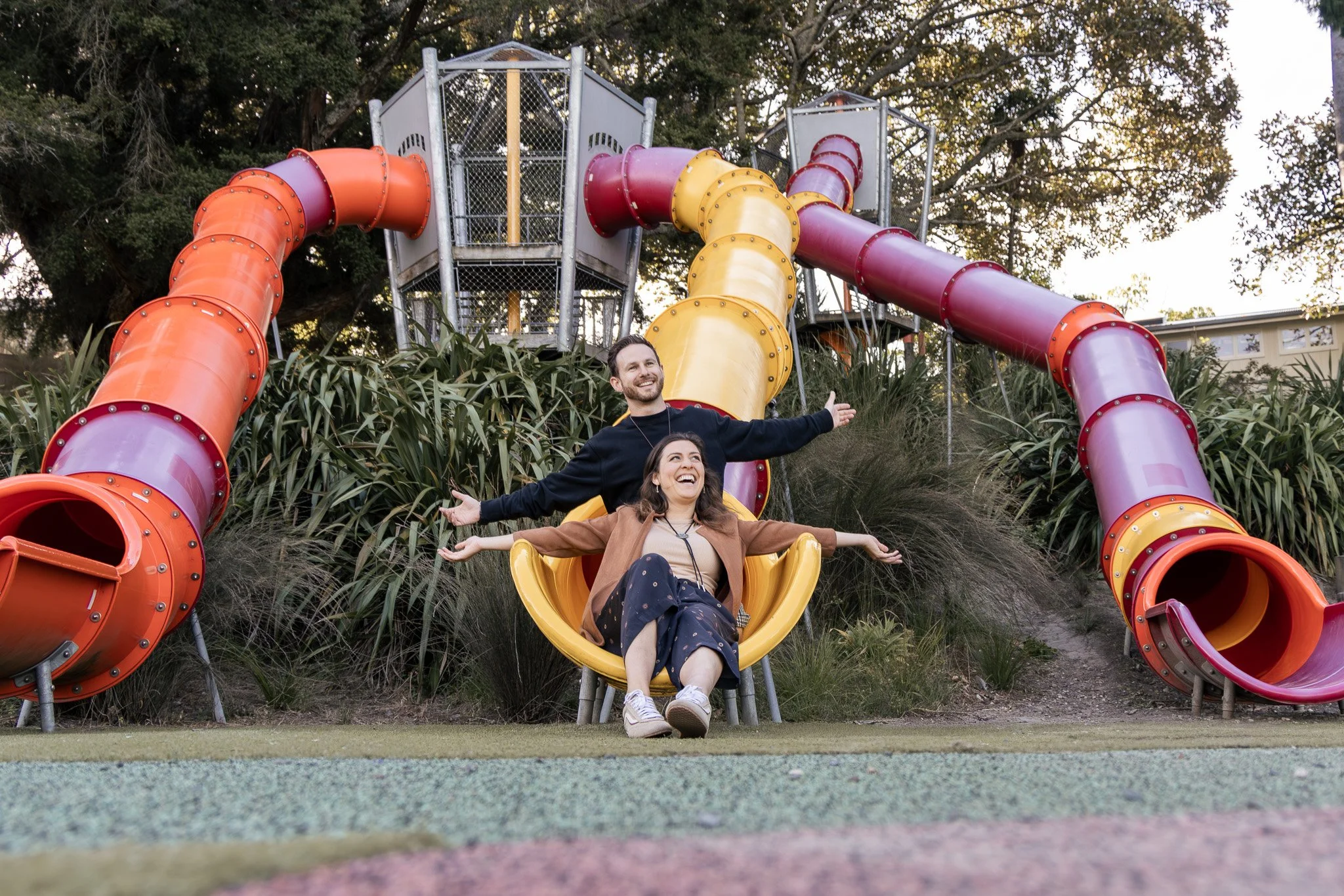 Two women and one man enjoying a yellow slide at a playground with colorful tubes, trees, and a building in the background.