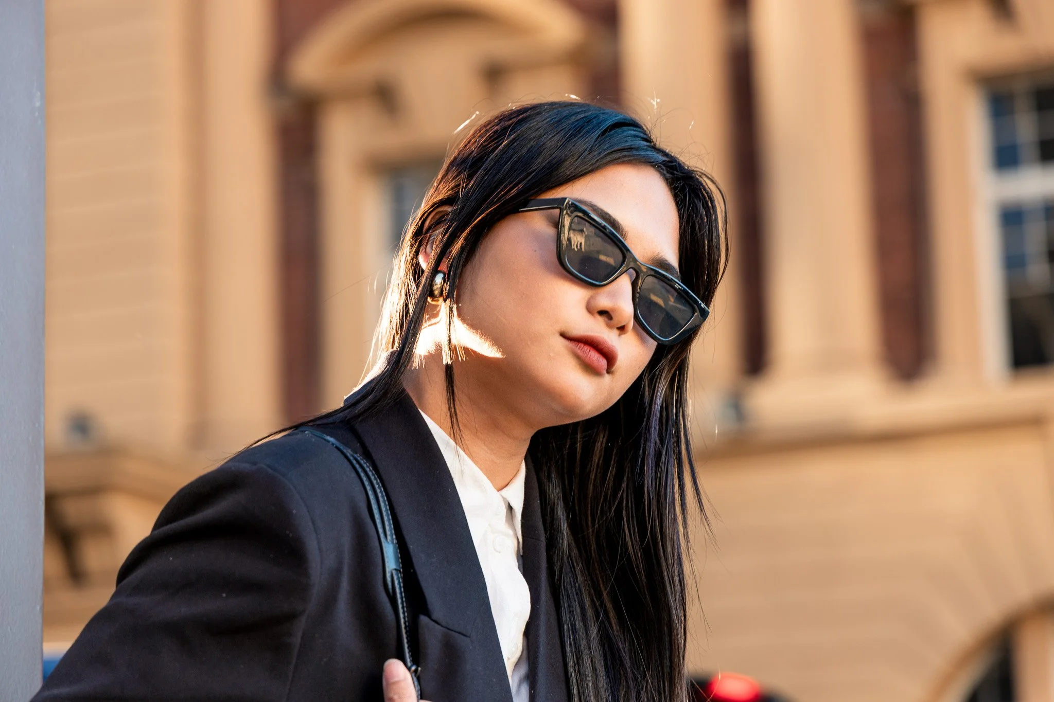 A woman with long black hair wearing black sunglasses, a black blazer, and a white shirt, standing outdoors with a building in the background.