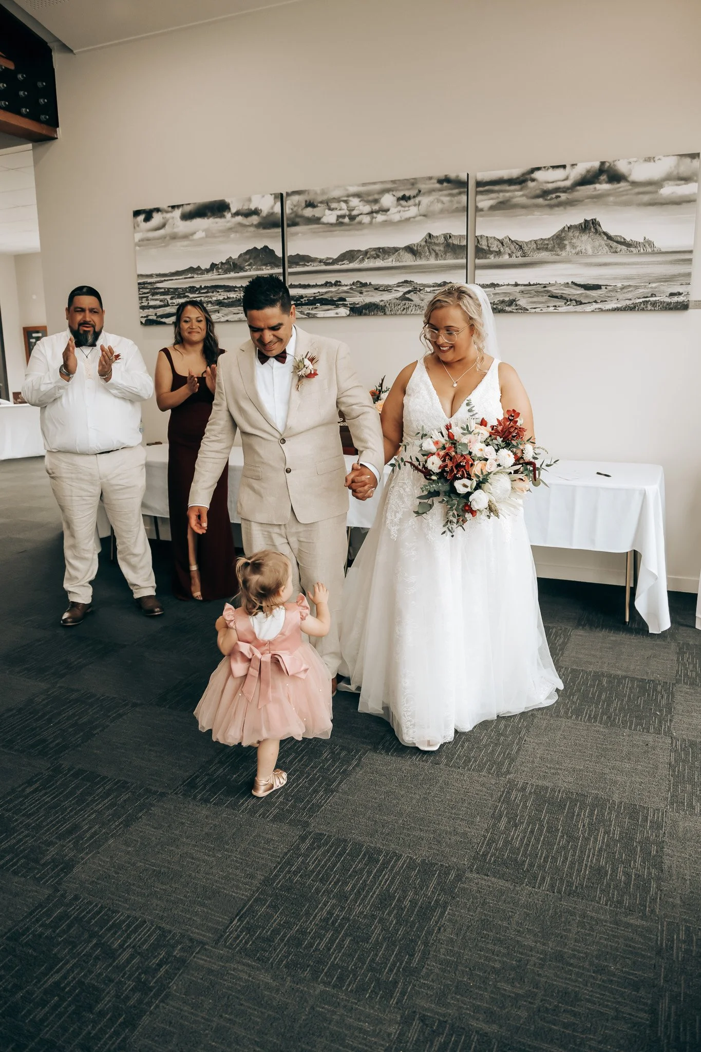 A wedding ceremony with four adults and a child. The bride is holding a bouquet, and the groom is holding her hand. The woman in a dark dress and the man in a white shirt are clapping in the background. The child, wearing a pink dress, is looking up 
