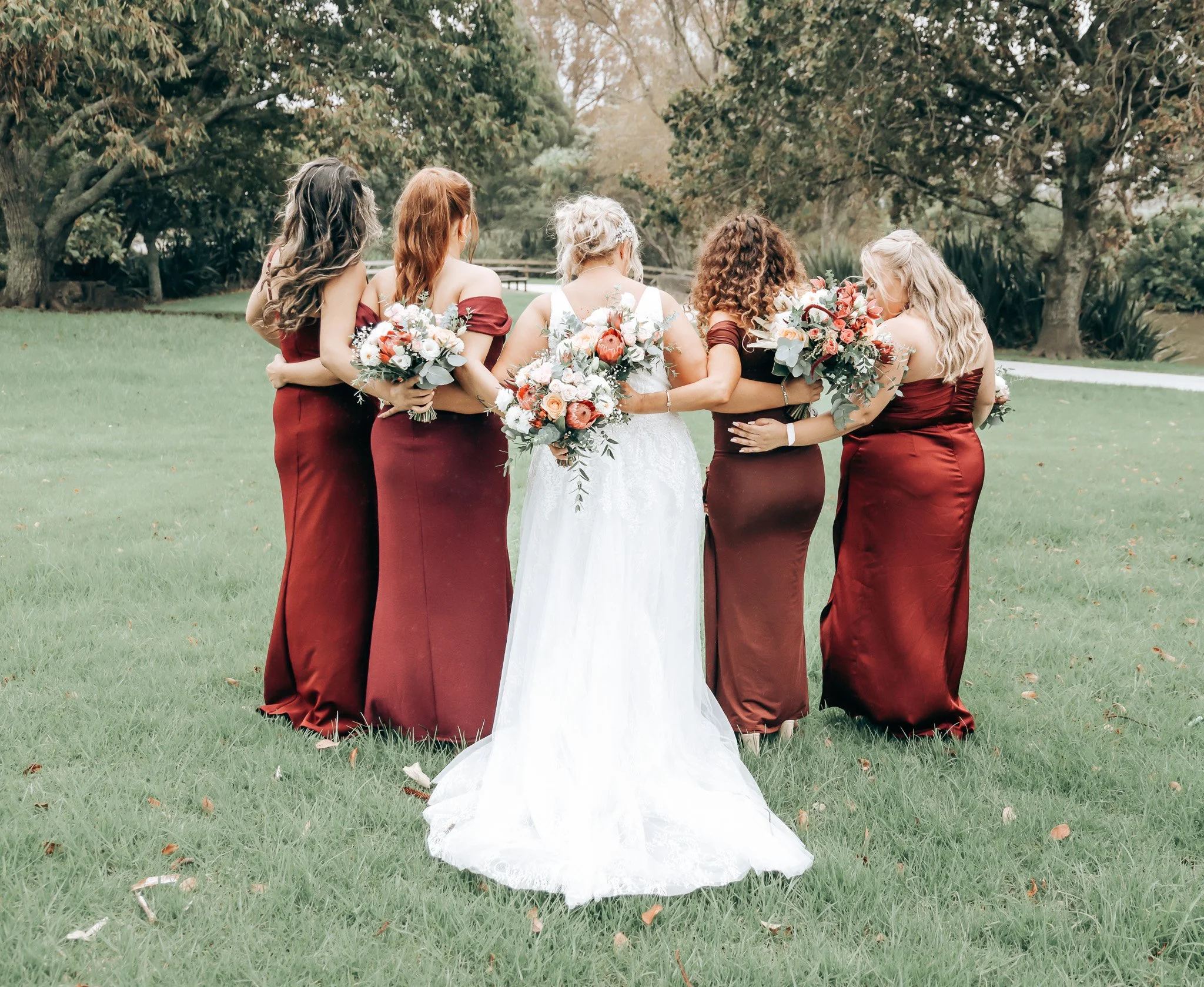 A bride and five bridesmaids standing in a grassy outdoor area with trees, with the bridesmaids wearing matching burgundy dresses and the bride in a white gown. All are holding bouquets of flowers.