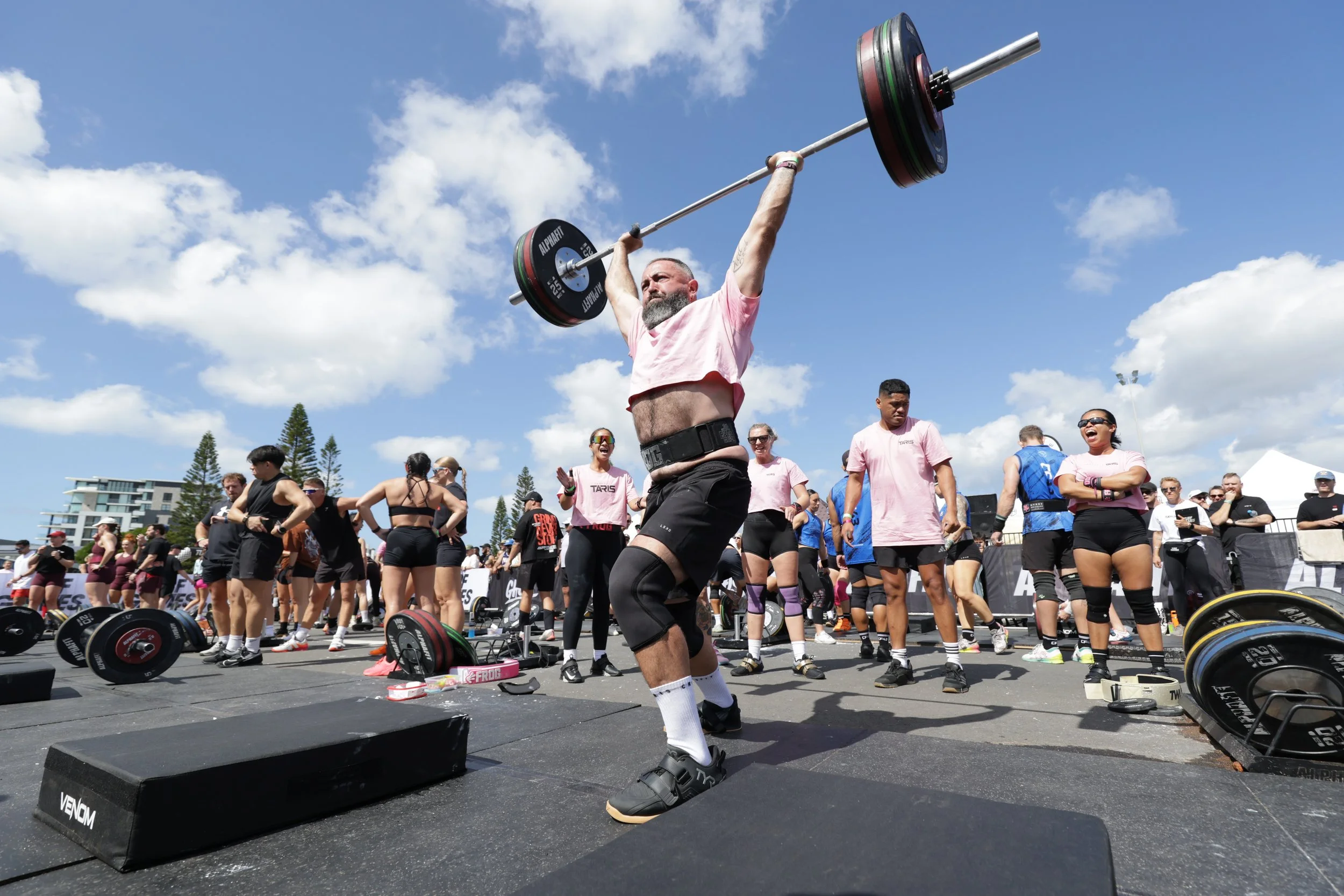 A man lifting a barbell overhead at an outdoor fitness event, surrounded by a group of people, some watching and others participating, with heavy weights on the ground and tents in the background under a blue sky with clouds.