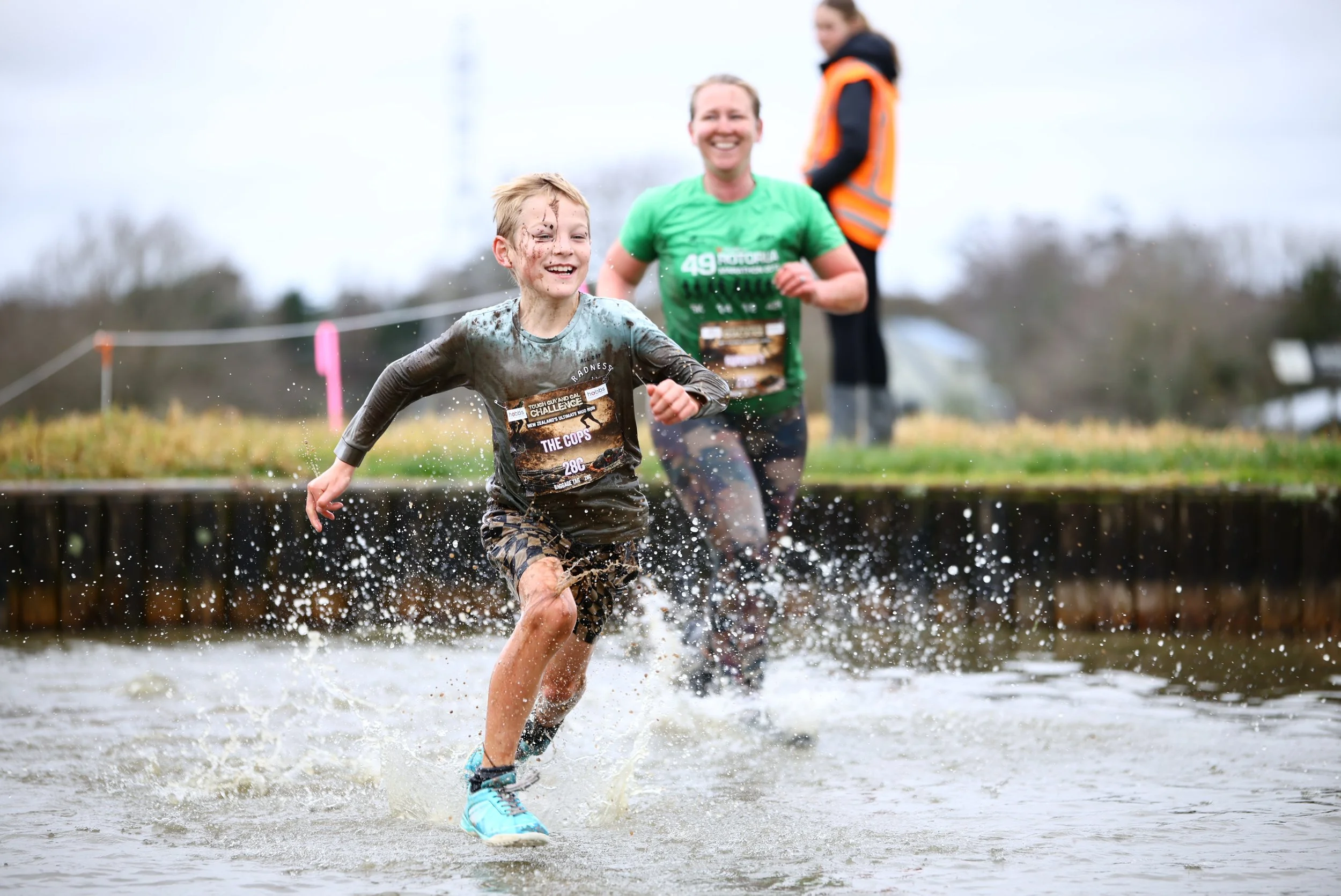 A young boy running through a muddy water obstacle during an outdoor mud run race, with a smiling man and a woman in the background.