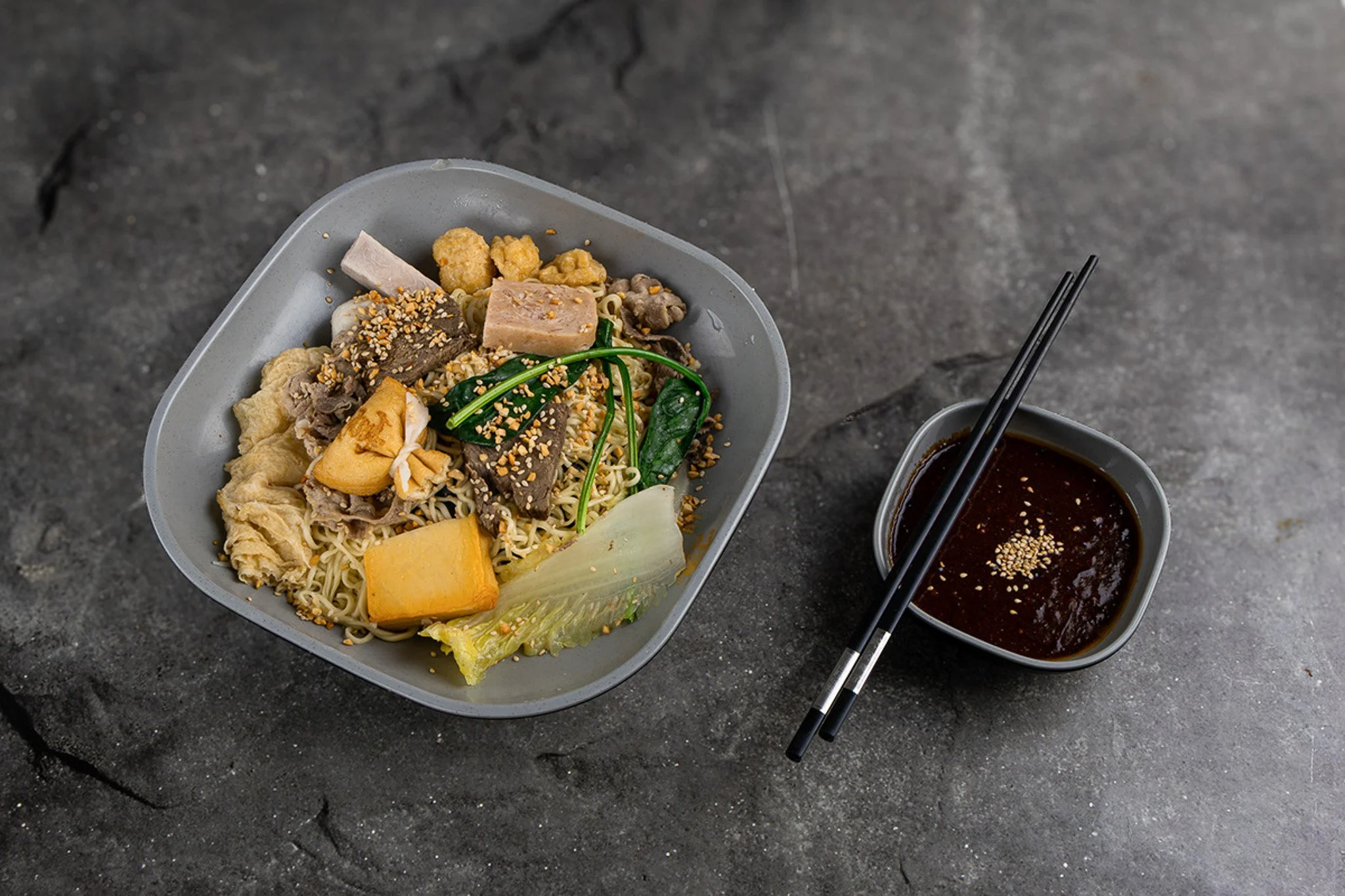 A takeout meal of Asian noodles with vegetables and meat in a gray bowl, with a side of dipping sauce in a black bowl with chopsticks on a dark, cracked surface.