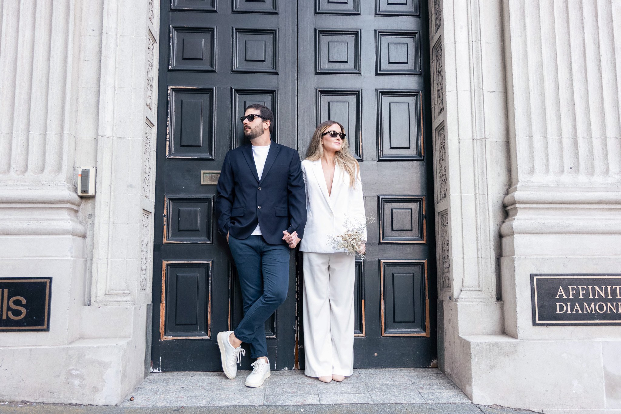 A man and woman stand outside a large black door with ornate stone columns on each side. The man wears a navy blazer, white shirt, sunglasses, and white sneakers, while the woman wears a white blazer, matching pants, sunglasses, and heels. They both 