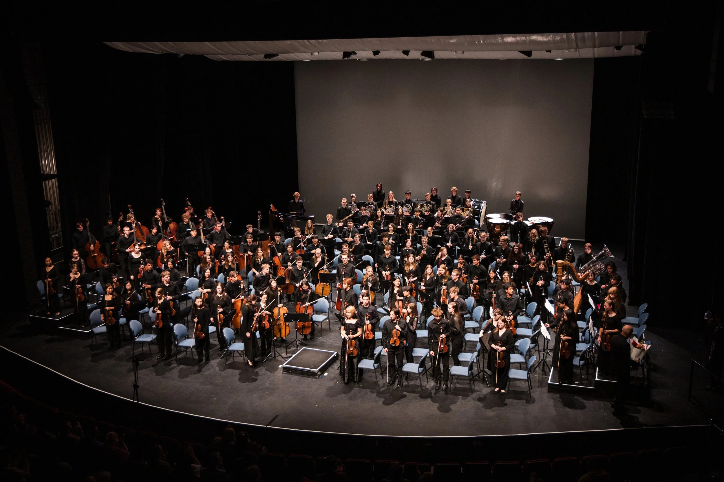 Orchestra of musicians on stage preparing for a performance with string, woodwind, brass, and percussion instruments.