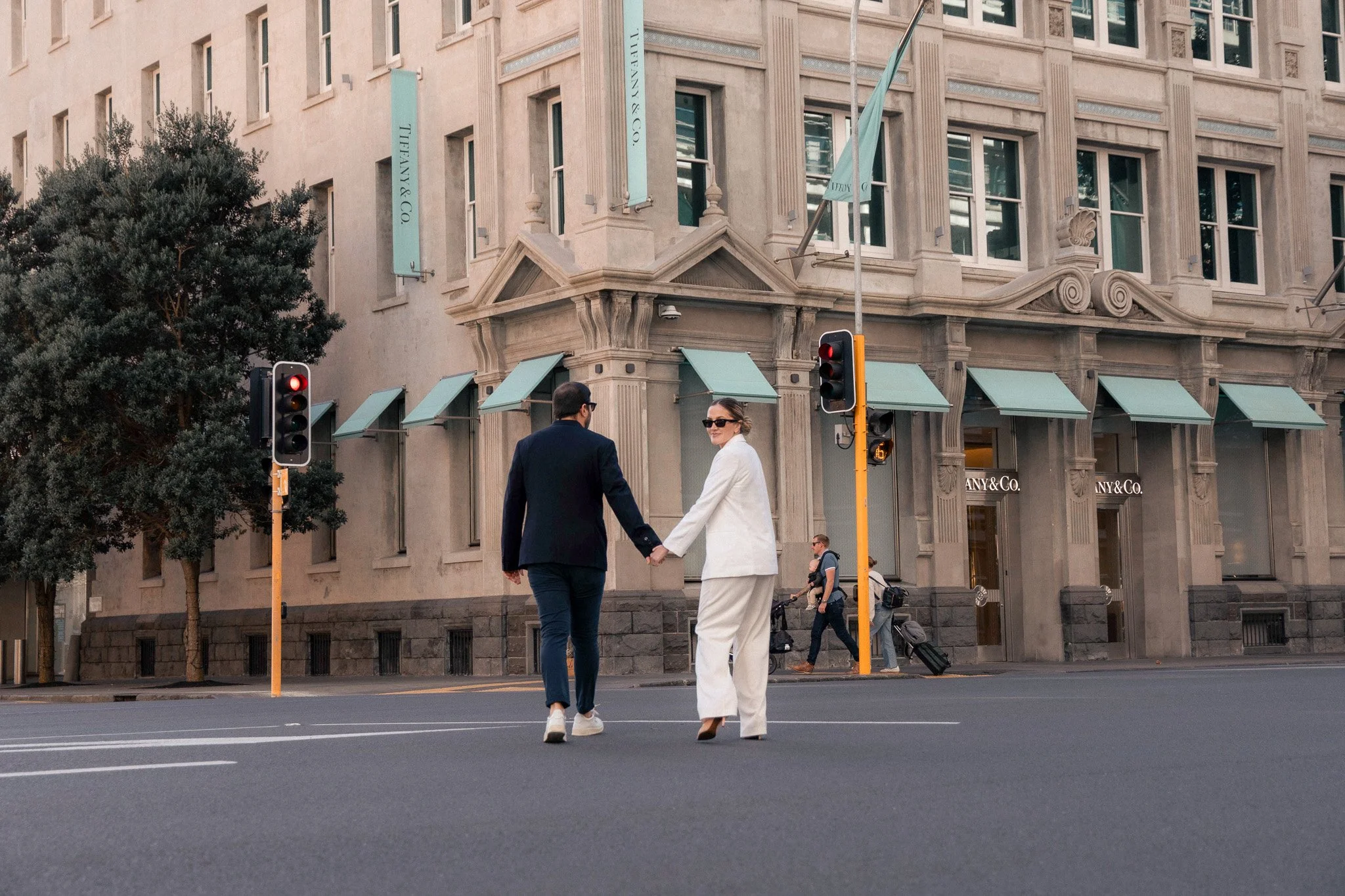 A man and woman holding hands and crossing the street in front of a Tiffany & Co. building, with traffic lights and pedestrians in the background.