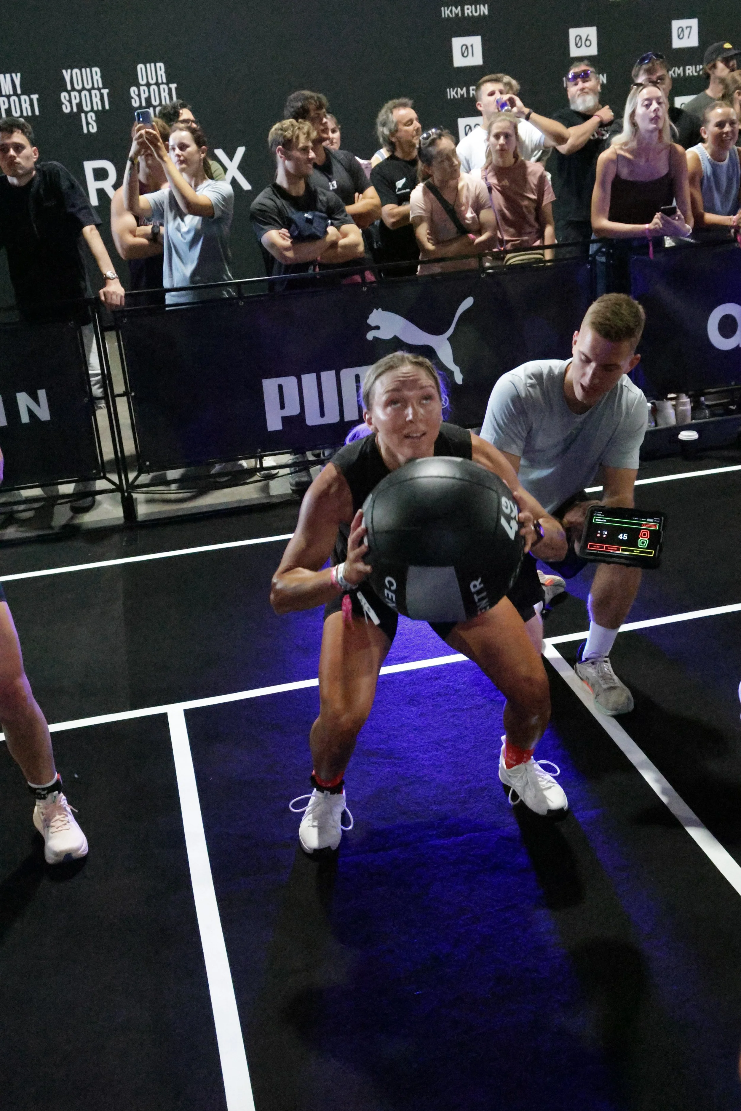 A woman is doing squats with a medicine ball at a fitness event, with a group of spectators watching behind a barricade. The background features branding and advertising for Puma, and the floor has court lines.