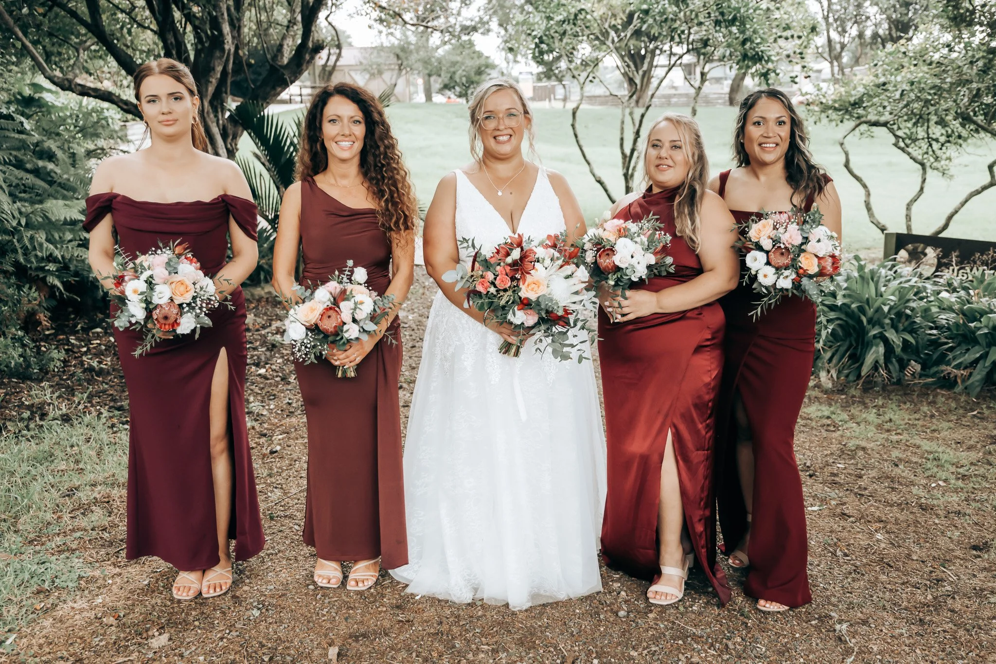 Group of five women in dresses, standing outdoors with trees in the background, holding bouquets, one in a white wedding dress, others in burgundy dresses.
