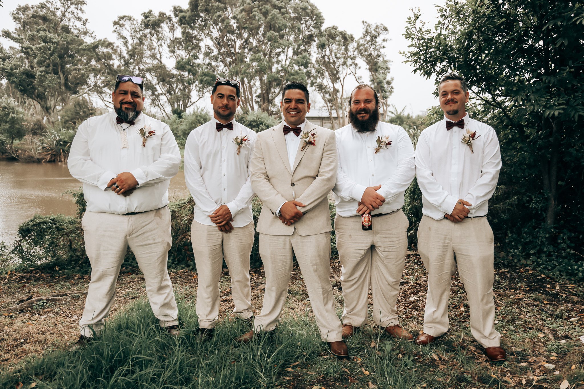 Group of five men in formal attire standing outdoors near a body of water, with trees in the background. Four men wear white shirts, brown bow ties, overalls, and boutonnières, while the central man wears a beige suit and a boutonnière.