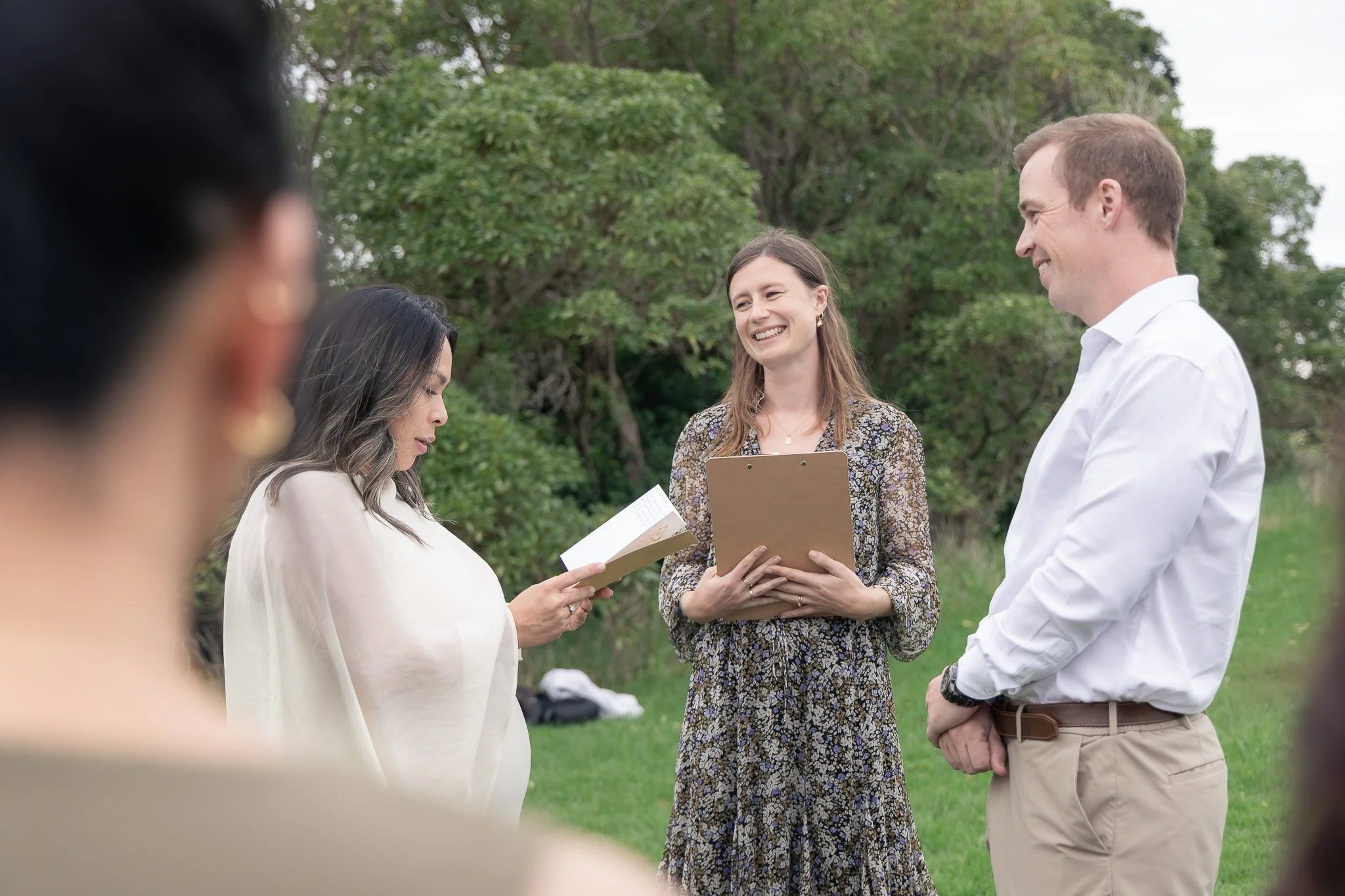 A group of people gathering outdoors under trees, engaged in a discussion or ceremony, with some looking at papers and a woman holding a clipboard smiling.