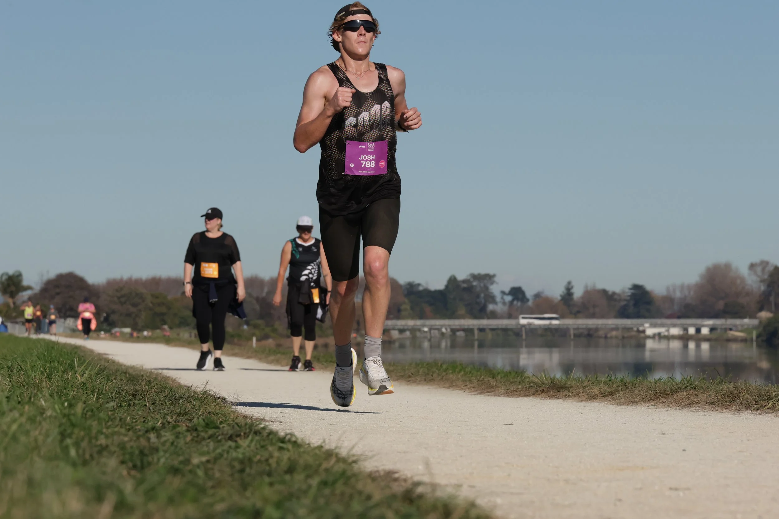 A person with short hair, wearing sunglasses, a tank top, and black shorts, running on a paved path near a river during a race or marathon. There are other runners in the background, some with race bibs. The weather is clear and sunny.