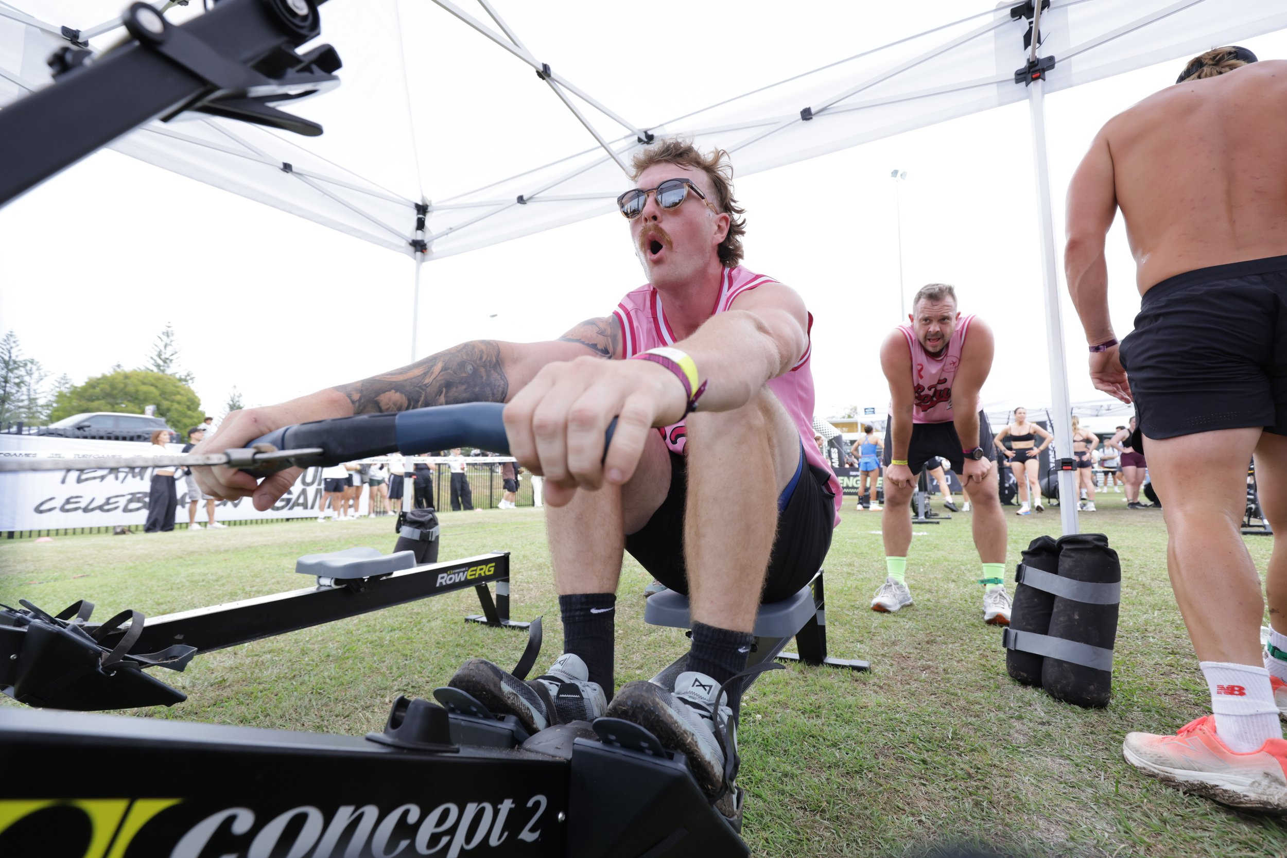 A man with sunglasses and a pink shirt participating in a rowing competition, leaning forward and pulling the oars, with other participants and spectators in the background under a white tent.