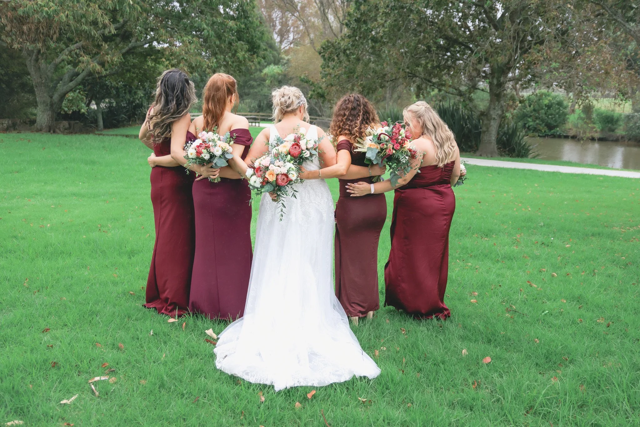 A bride in a white wedding dress, surrounded by five bridesmaids in matching burgundy dresses, holding bouquets of mixed flowers, standing outdoors on a grassy area near a pond with trees in the background.