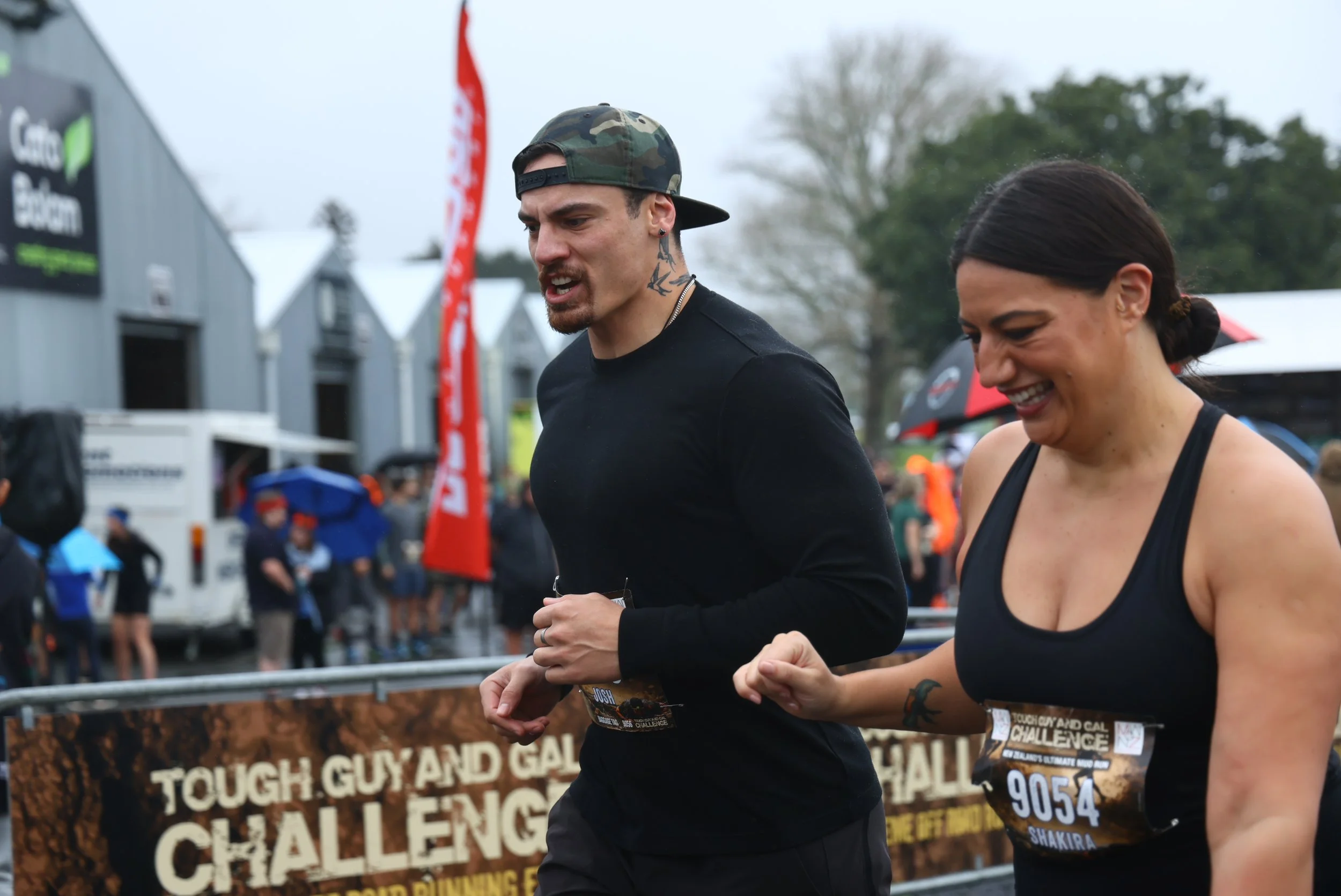 Two people participate in the Touch Guy and Gal Challenge outdoors during cloudy weather, with event banners and a crowd in the background.