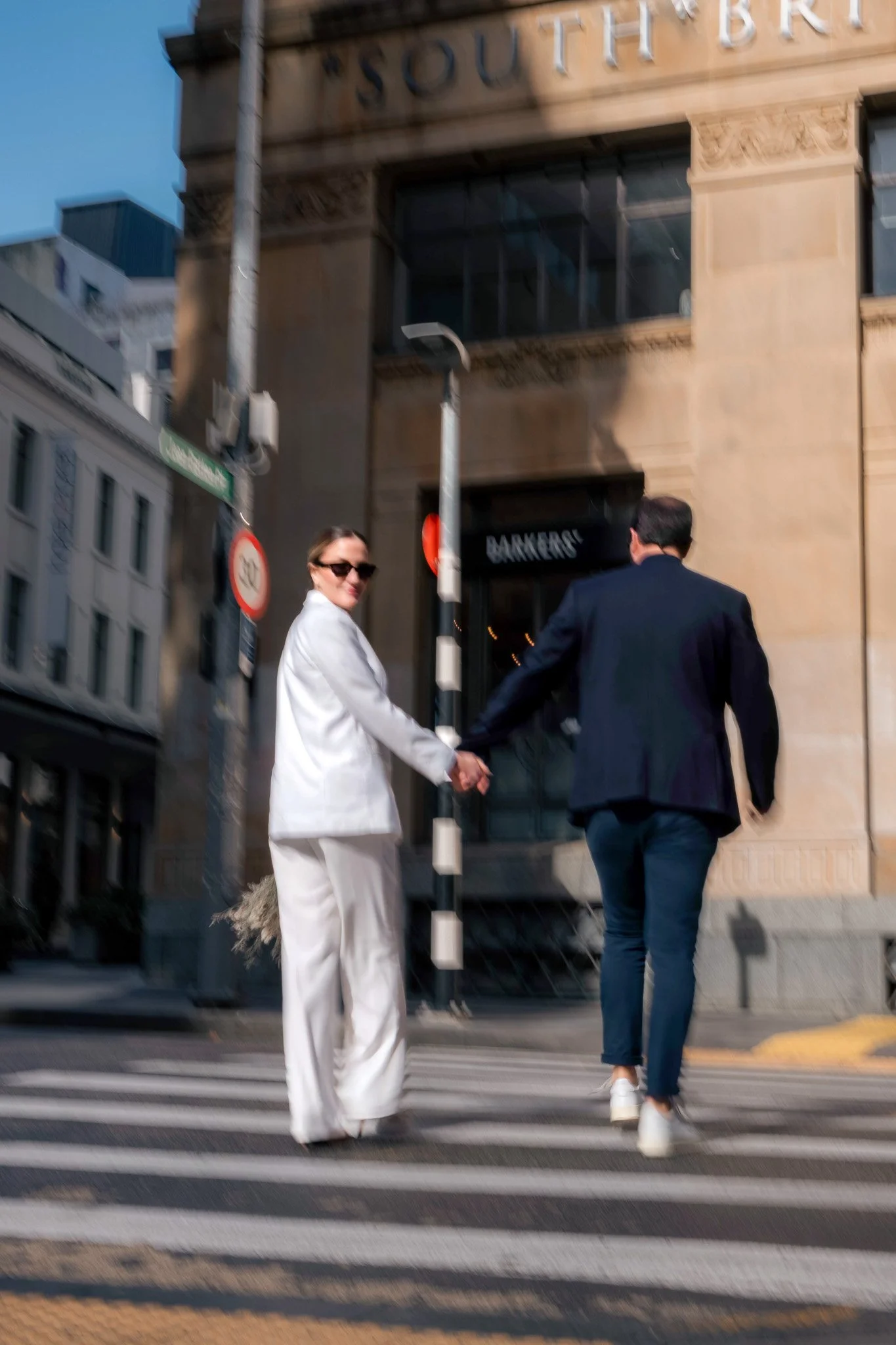 A woman in a white suit and sunglasses holding hands with a man in a dark blazer walking across a city street at a crosswalk, with buildings and street signs in the background.
