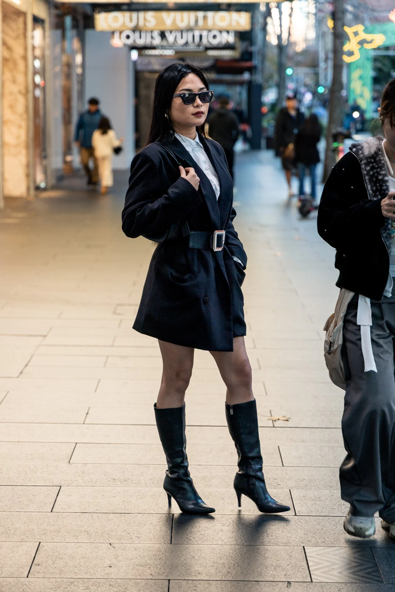 A woman wearing sunglasses, a black blazer, a white shirt, and high-heeled boots standing on a city sidewalk.