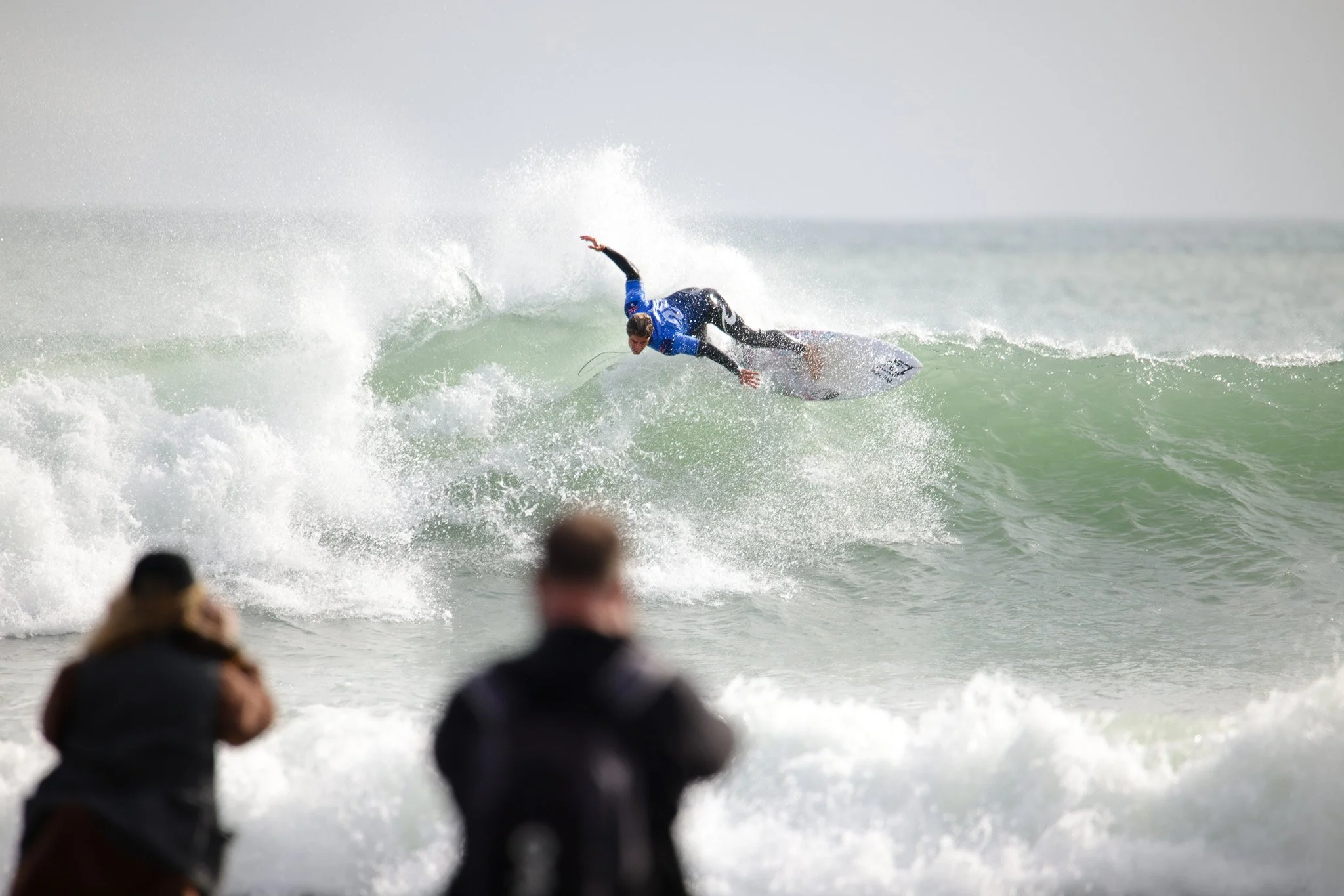 A surfer dressed in a blue wetsuit riding a wave on a surfboard, with spectators watching from the shore.