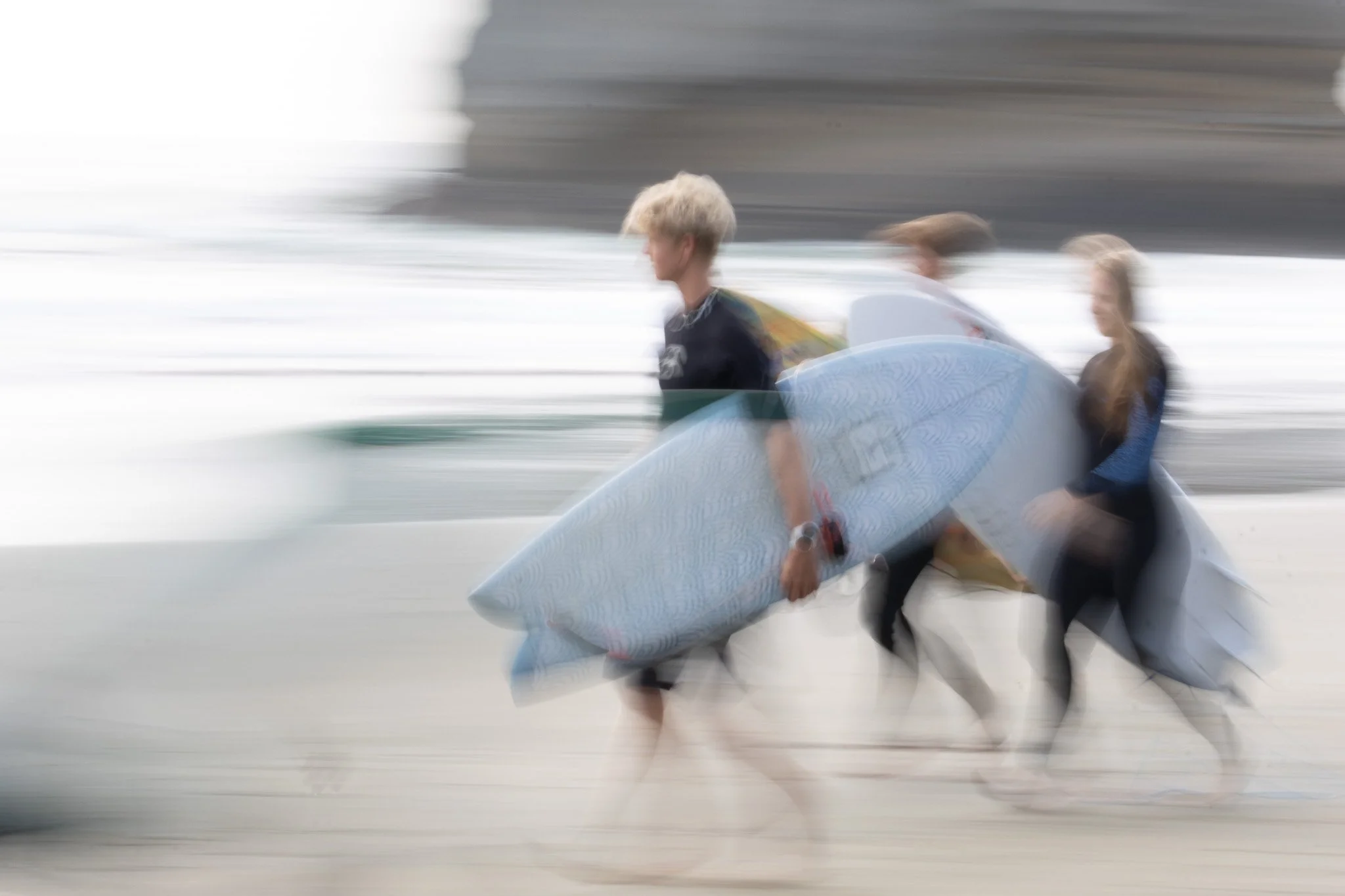 Two surfers walking along a beach with surfboards, captured with motion blur.