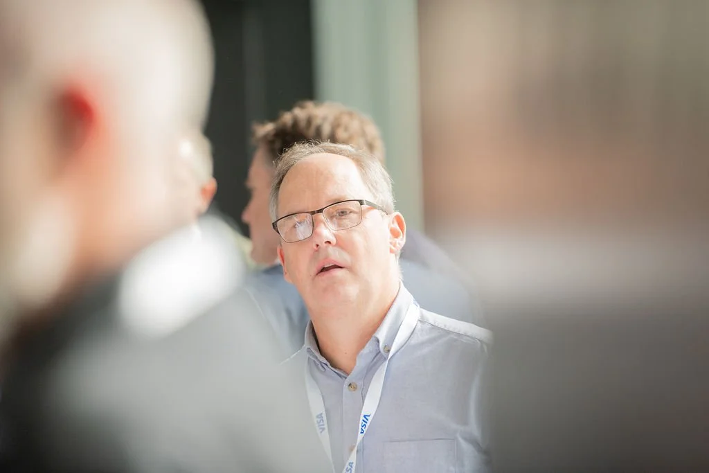 A man with glasses and a lanyard appears to be talking or listening during a meeting, with blurred people around him.