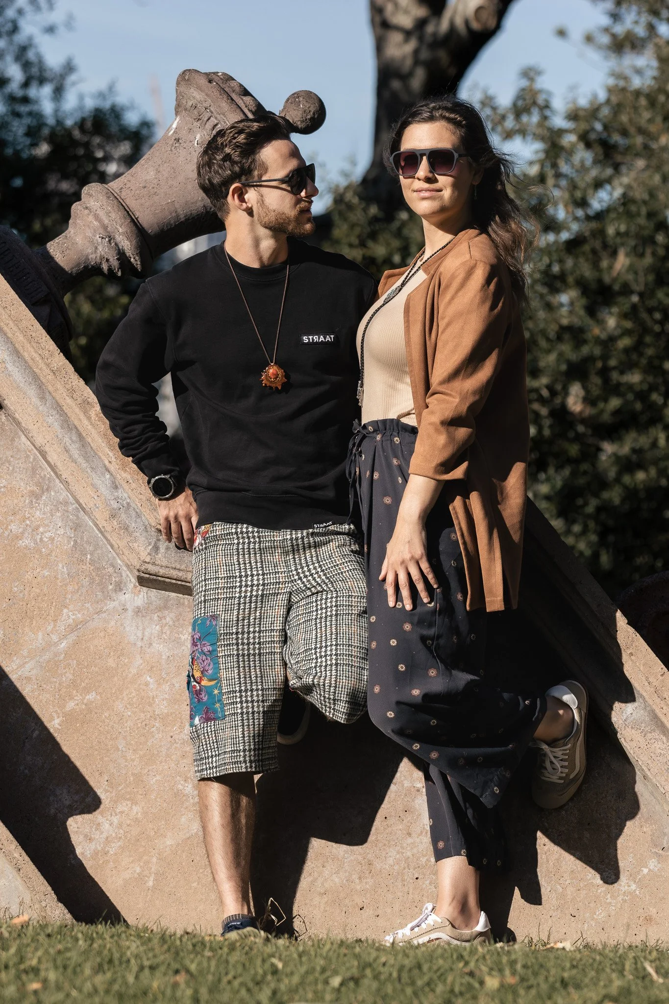 Two people, a man and a woman, stand close to each other outdoors, with a large tree and a cloudy sky in the background. They are both wearing sunglasses and casual clothing, with the man dressed in a black sweater and checkered shorts, and the woman