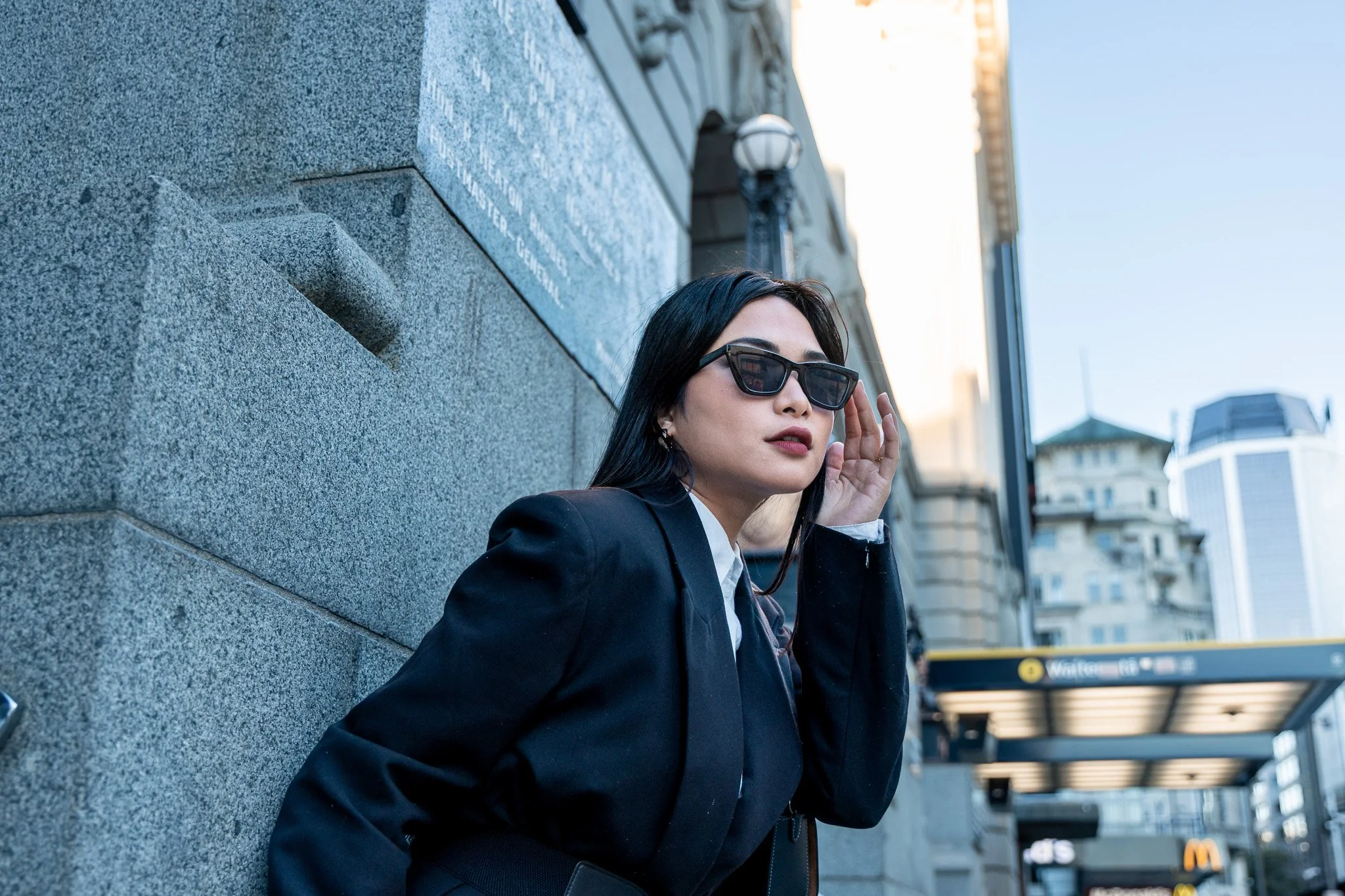 A woman with black hair, wearing sunglasses and a dark blazer, is touching her ear while leaning against a gray stone building on a city street in the late afternoon.