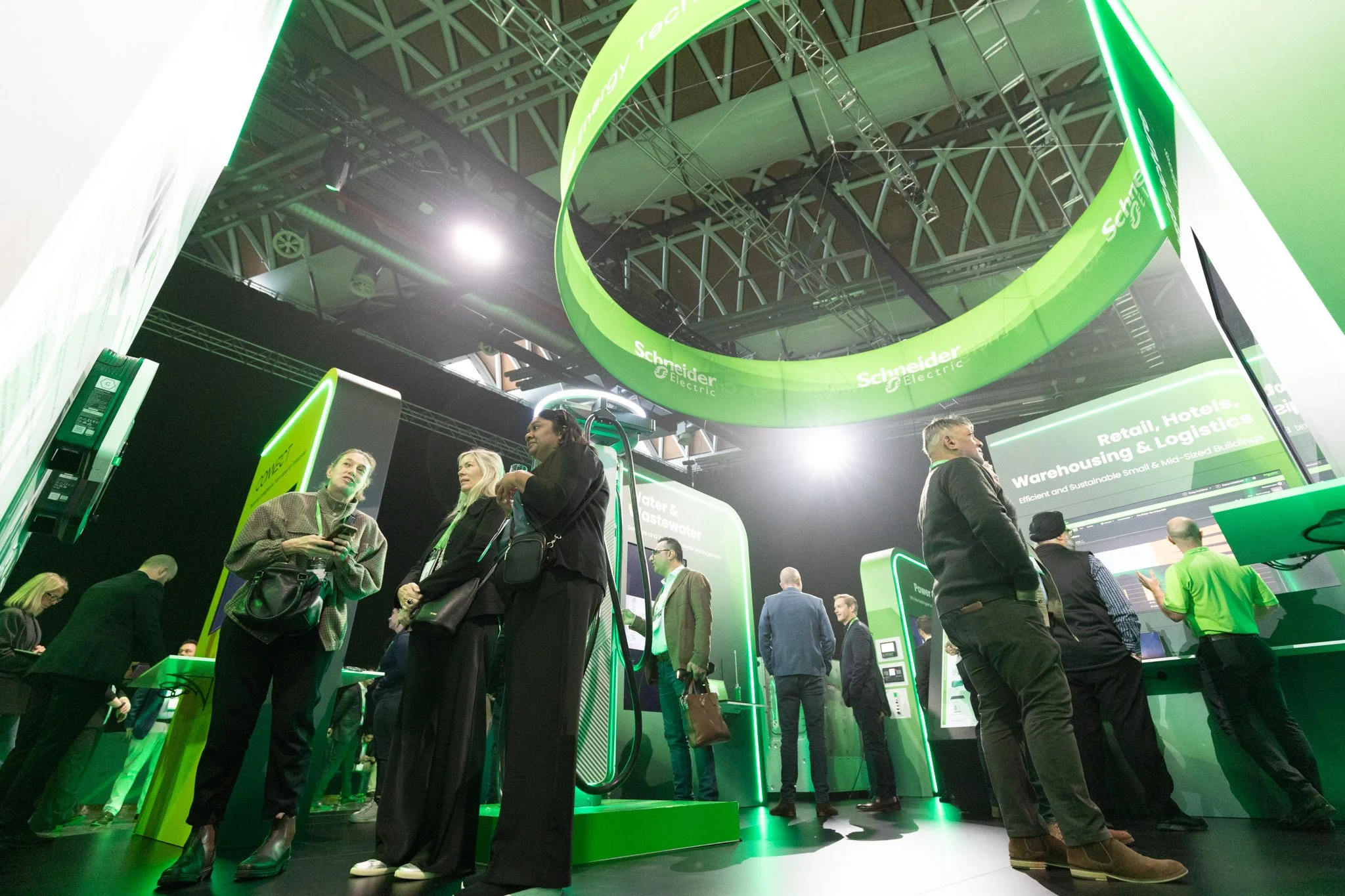 People gathering at an exhibition booth for Schneider Electric, displaying green and black technology equipment and digital screens, with a large circular green banner overhead.