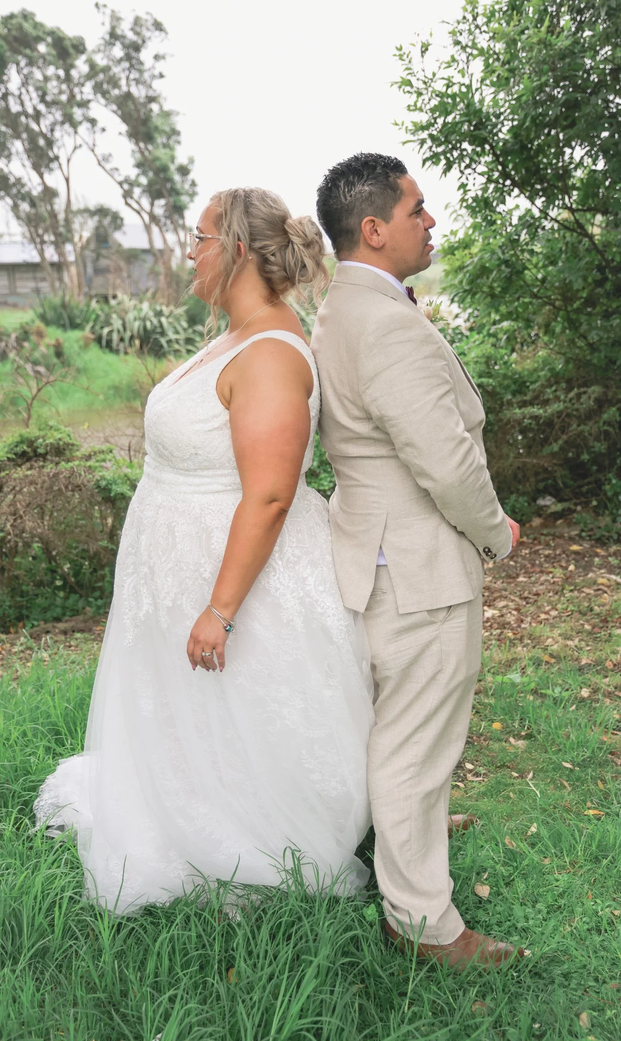 A couple dressed in wedding attire standing back to back outdoors in a green garden with trees and grass.