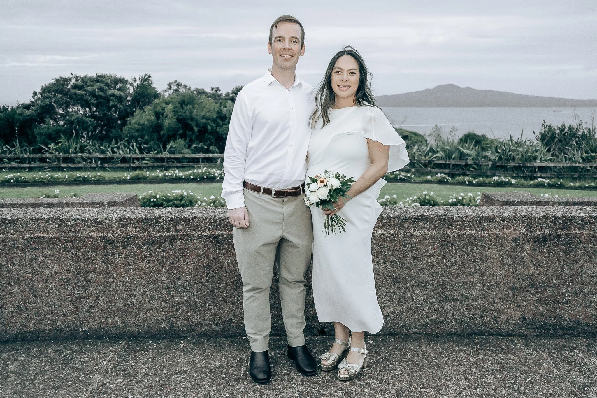 A couple standing together outdoors on a cloudy day, with a scenic landscape and water in the background. The woman is holding a bouquet of flowers, and both are dressed in white and light-colored clothing.