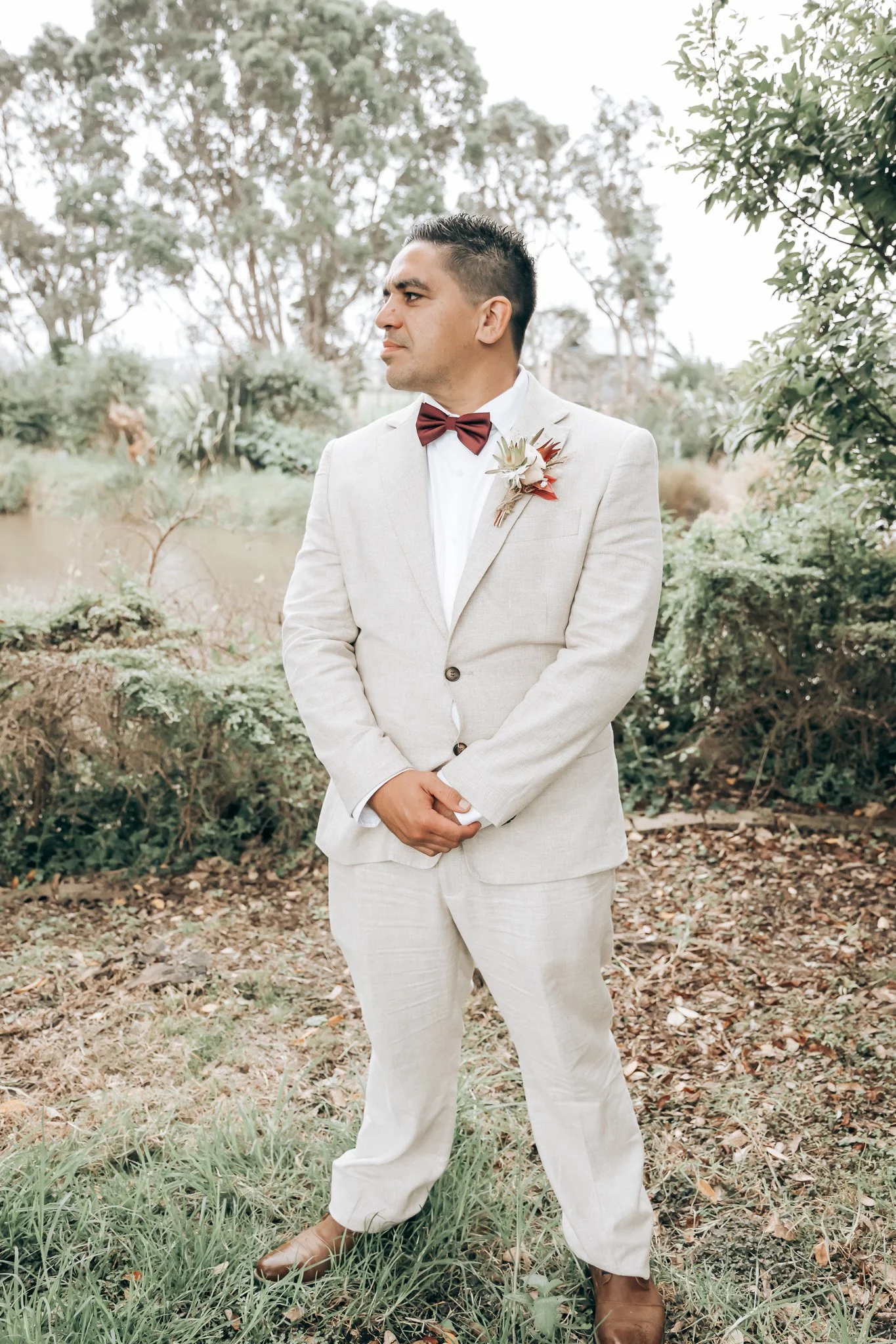 A man in a light-colored suit and brown shoes standing outdoors near a small body of water with trees in the background, wearing a white shirt and a brown bowtie, with a boutonniere on his lapel.