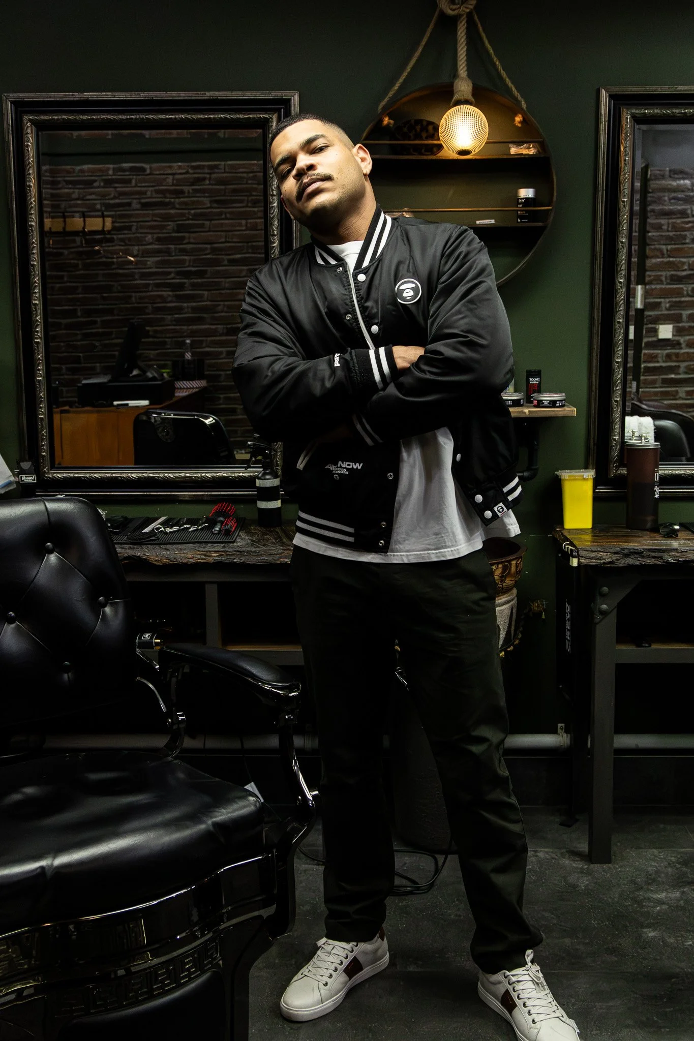 A man stands in a barbershop or salon with arms crossed, wearing a black varsity jacket, white sneakers, and black pants. The background includes mirrors, brick walls, and barber tools, with warm lighting from a hanging lamp.