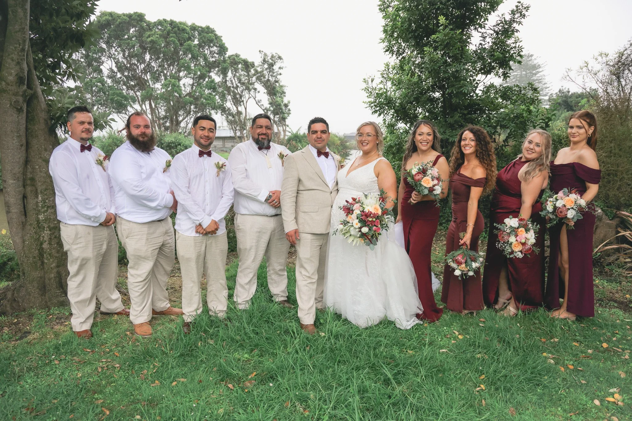 A group of ten people, including a bride and groom, standing outdoors on grass surrounded by trees, at a wedding celebration, with joyful expressions and holding bouquets.