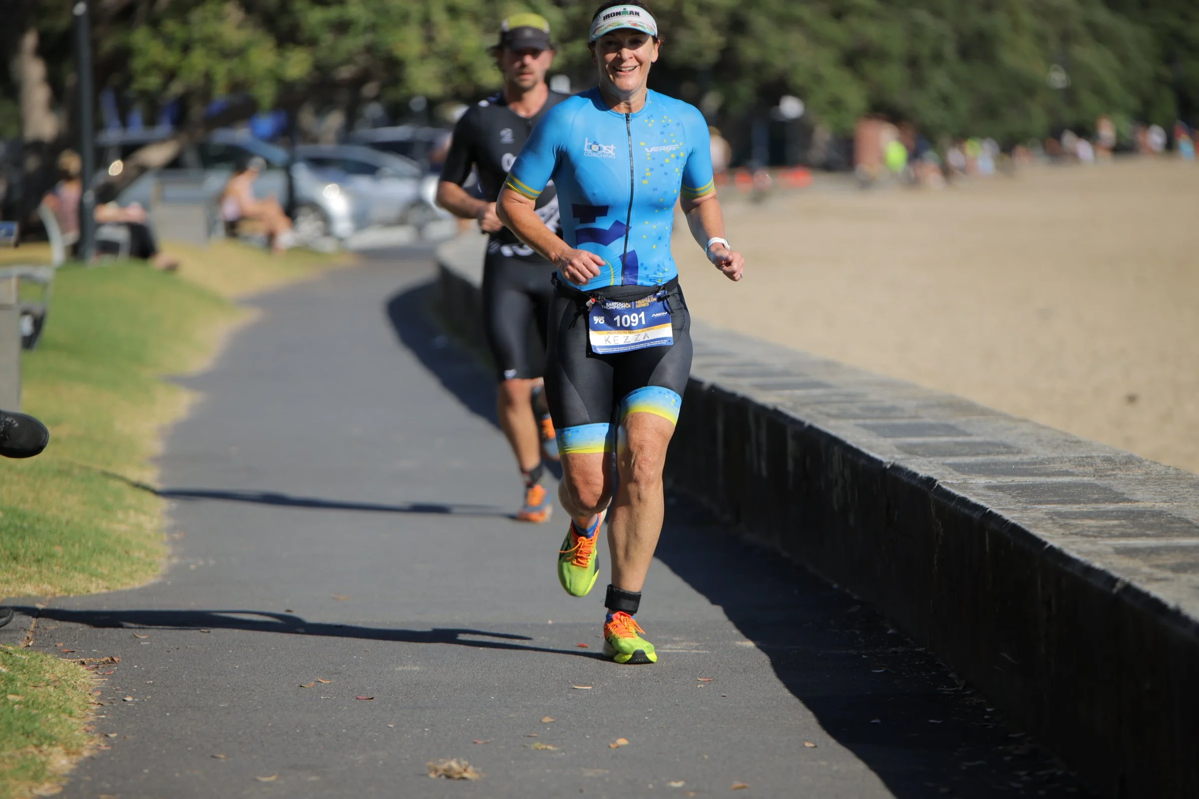 A woman in a blue and black athletic outfit running outdoors on a pathway, with a race bib labeled 'KEZZA' and the number 1091, smiling as she participates in a race or marathon. There are trees and parked cars in the background, and a man running be